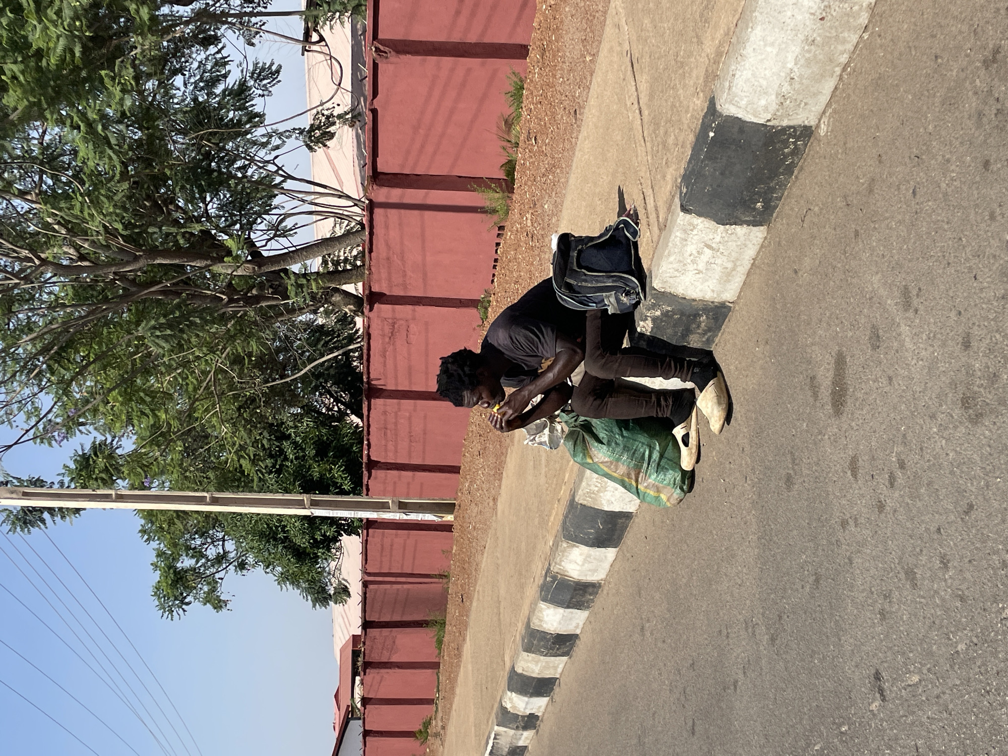 A lone individual is captured in a daylight scene, crouching on the edge of a paved sidewalk next to a road. The person has dark skin and dark, possibly braided or curly, hair, and is wearing a dark short-sleeved top and dark trousers with light-colored shoes. They appear to be engaged in eating or preparing food, holding a small yellowish item close to their mouth.
Next to the person, a black backpack rests against the curb, and a green and white striped woven sack is on the ground near their feet. The sidewalk is a light brown, paved surface, bordered by a concrete curb painted with distinct alternating black and white stripes, separating it from a grey asphalt road.
In the background, a reddish-brown wall stretches horizontally across the scene, likely marking a property boundary. Lush green trees with visible branches and foliage rise above and behind this wall, casting some dappled shade. Above the trees and wall, a clear, bright blue sky is visible, indicating a sunny day. Power lines are faintly discernible against the sky in the upper left portion of the image. The overall setting appears to be an urban or semi-urban outdoor environment. No visible text is present.