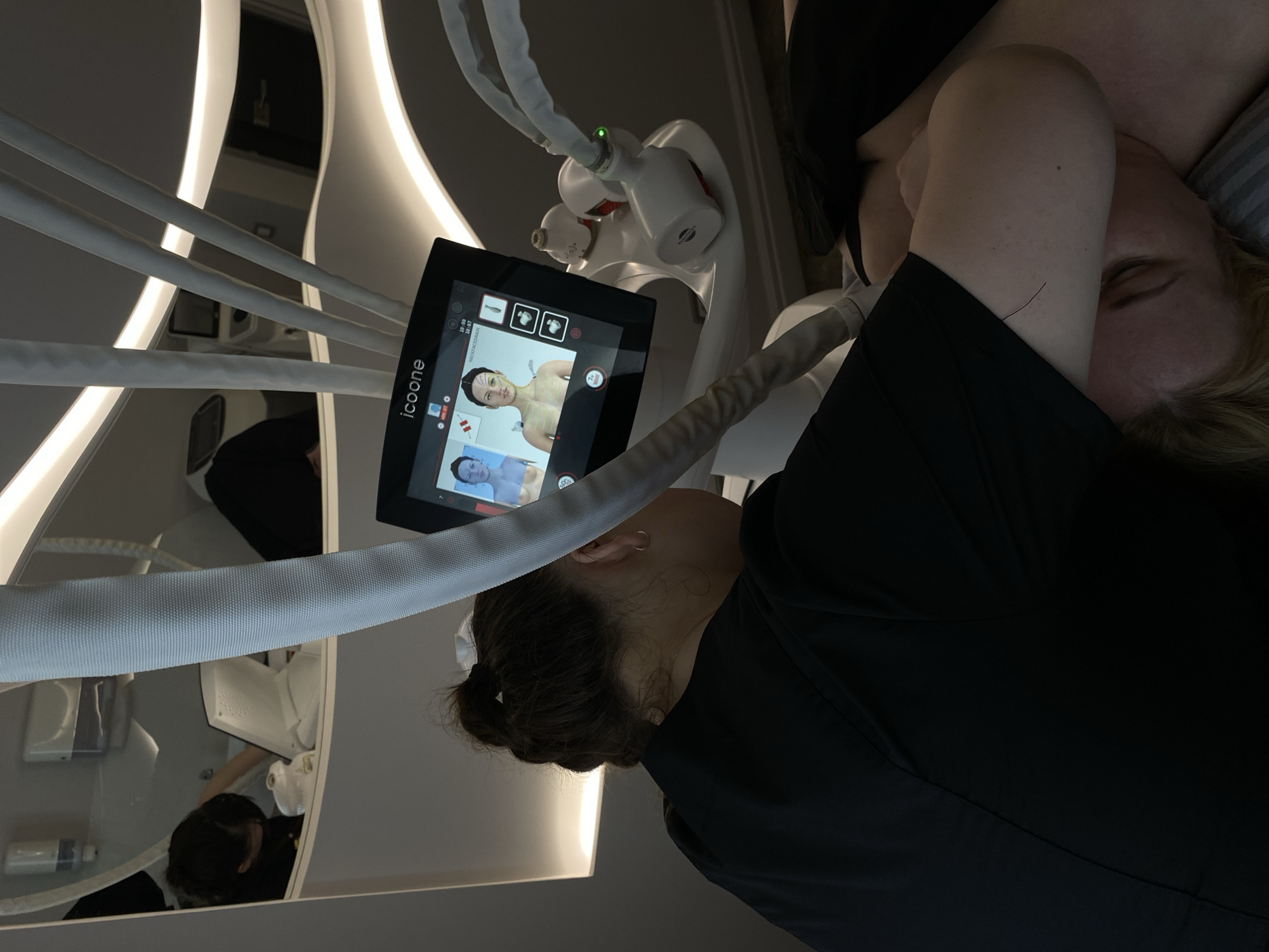 A woman is lying down, partially obscured by a black shirt. An Icone medical device with a touchscreen displaying treatment options is positioned beside her. A person wearing black is operating the device. The scene appears to be in a medical spa or cosmetic treatment facility. The exact location in Votkinsk, Russia, cannot be determined from the image.