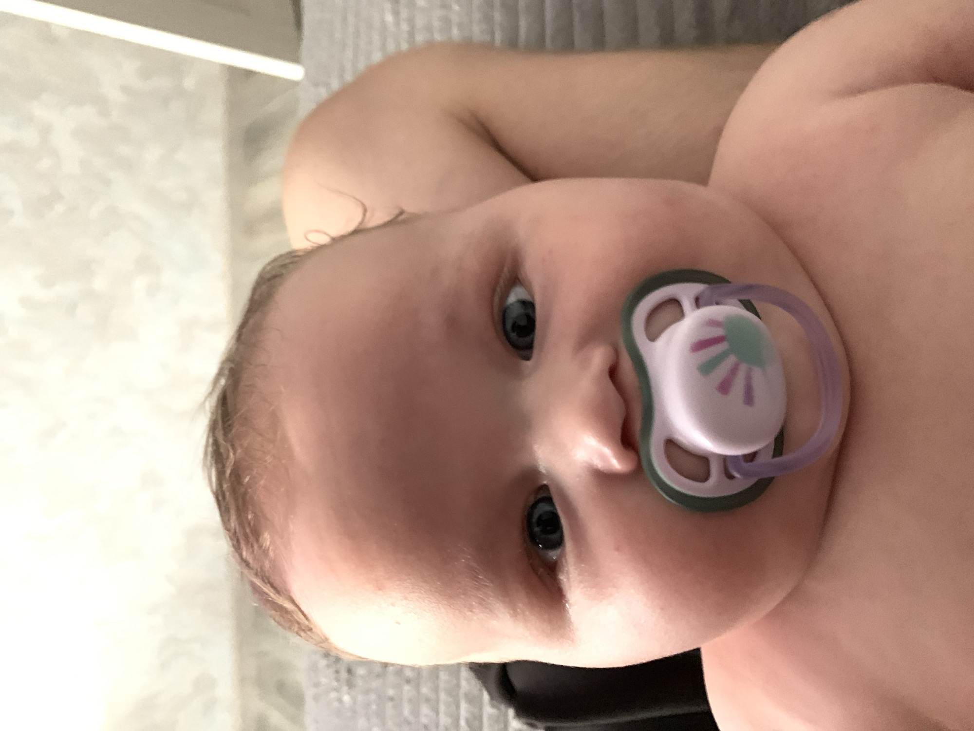 This is a close-up, rotated photograph of a baby's face, featuring a pacifier. The baby, whose face is the primary subject, has light skin, blue eyes, and fine, light brown hair. A pink and purple pacifier is in the baby's mouth. The background is out of focus, but appears to be a domestic setting with a textured surface, possibly a blanket or upholstered furniture, in shades of gray. There is also a bright horizontal light source visible in the upper left portion of the image. The lighting suggests it could be daytime, possibly indoors. No text is visible in the image. The baby's expression is neutral.