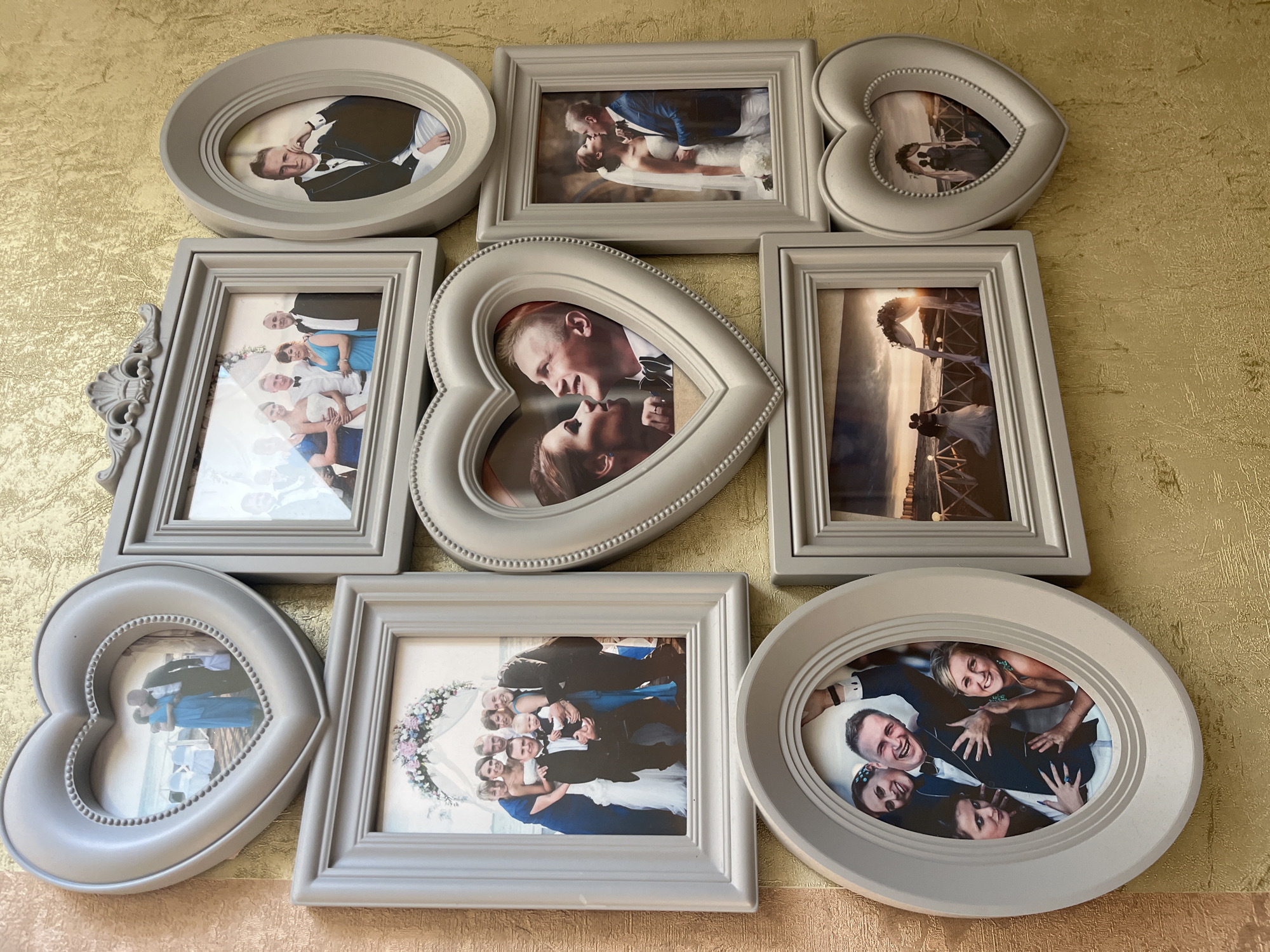 A collection of framed photographs is displayed on a textured, golden-yellow wall. The frames are predominantly a light gray color and come in various shapes, including hearts, ovals, and rectangles.
In the top left, a heart-shaped frame contains a photograph of a couple embracing on a beach, with white chairs and an archway in the background.
To the right of the heart-shaped frame, a rectangular frame holds an image of a wedding group, including a bride in a white dress, groomsmen in tuxedos, and a woman in a blue dress. A baby is held by one of the groomsmen.
To the right of that, another oval frame displays a man in a tuxedo, with his arm resting on his knee.
Below the first heart-shaped frame, a rectangular frame shows a wedding ceremony archway decorated with flowers, with a couple silhouetted against a sunset.
To the right of this, a larger heart-shaped frame contains a close-up photograph of a couple kissing, with the man looking up at the woman.
Below the wedding group photo, an oval frame captures a man being hugged by three women, all smiling and appearing joyful.
To the right of the oval frame with the group, another rectangular frame shows a wedding couple in front of an archway by the water at sunset.
In the bottom right corner, a small heart-shaped frame contains a silhouette of a couple standing near a wedding archway.
The scene suggests a home or a place where wedding memories are displayed. The location is not specified in the image itself.