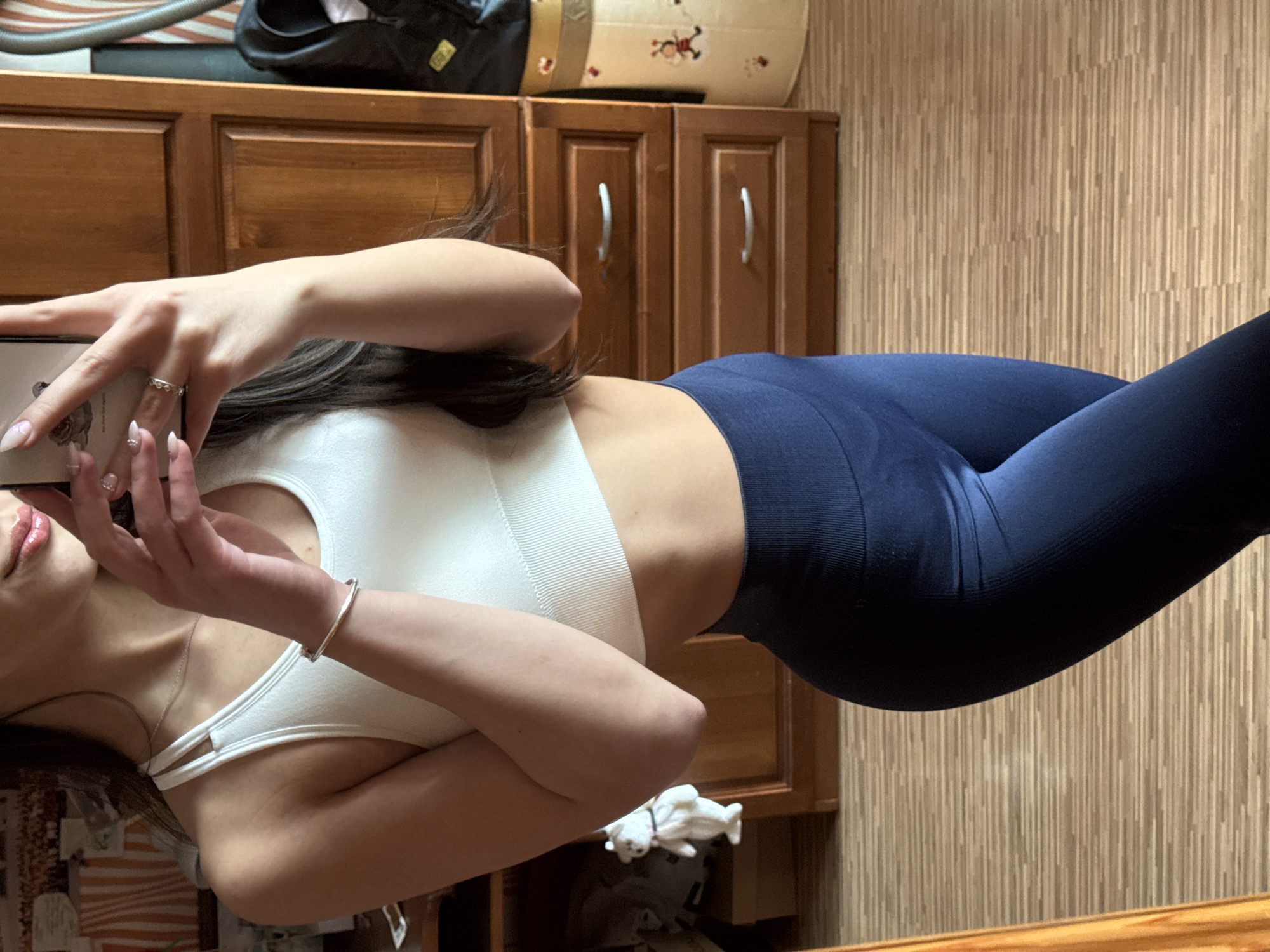 A woman wearing a white sports bra and blue leggings is taking a selfie. She is indoors with wooden cabinets and a patterned wallpaper visible in the background. The location is Unknown, Unknown.