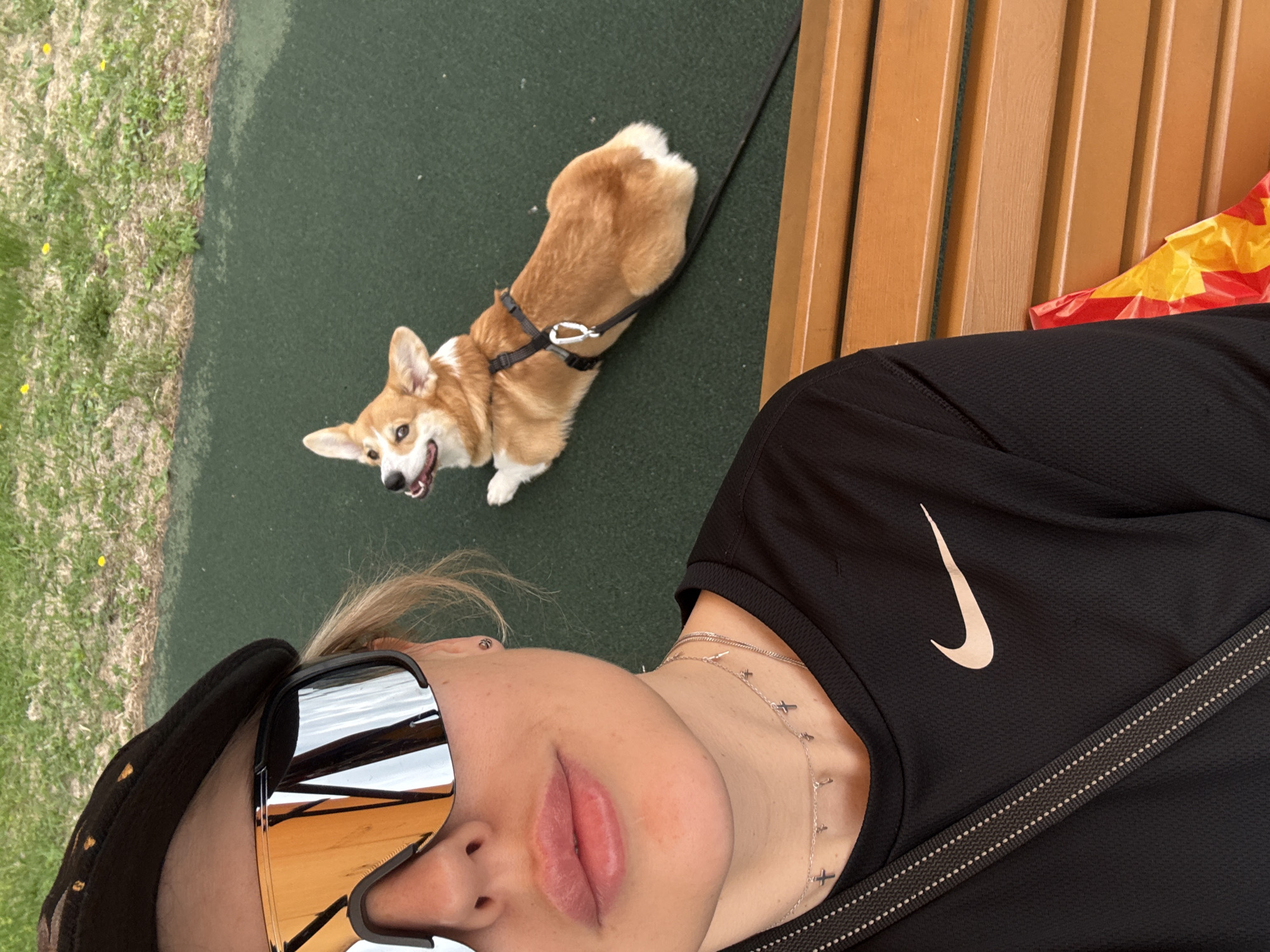 A selfie taken outdoors shows a light-skinned person with blonde hair, wearing dark sunglasses and a dark cap, and a black Nike athletic shirt, lying on a brown wooden bench. A necklace with cross charms is visible on their neck. A corgi dog on a black leash sits on a green surface, presumably grass or artificial turf, to the left of the bench. The dog is wearing a harness. The background suggests an outdoor recreational area, potentially in Krasnoyarsk, Russia. Part of a yellow and red object is visible on the bench next to the person. The photo has been rotated.