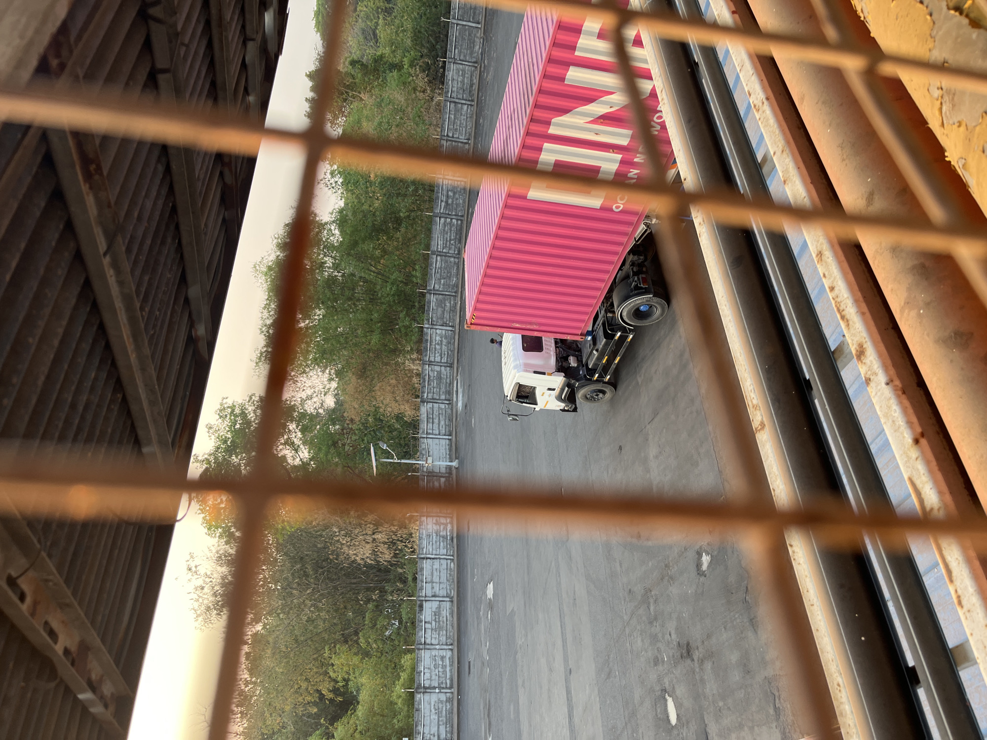 An outdoor, daytime, high-angle shot captures a large, pink shipping container atop a white truck parked on a gray asphalt lot. The container is emblazoned with white lettering that reads "ONE OCEAN NETWORK" and "ONE". The truck is positioned to the left of the container. In the background, a low, concrete wall with metal fencing runs horizontally, separating the lot from a line of trees. A solar-powered street lamp stands near the center of the lot. The scene is viewed through an orange, metal grate, which partially obscures the foreground. The sky above is a pale orange, suggesting either dawn or dusk. The image is taken from an elevated vantage point, possibly a building or structure overlooking the lot. The location is identified as Pelang, Indonesia.