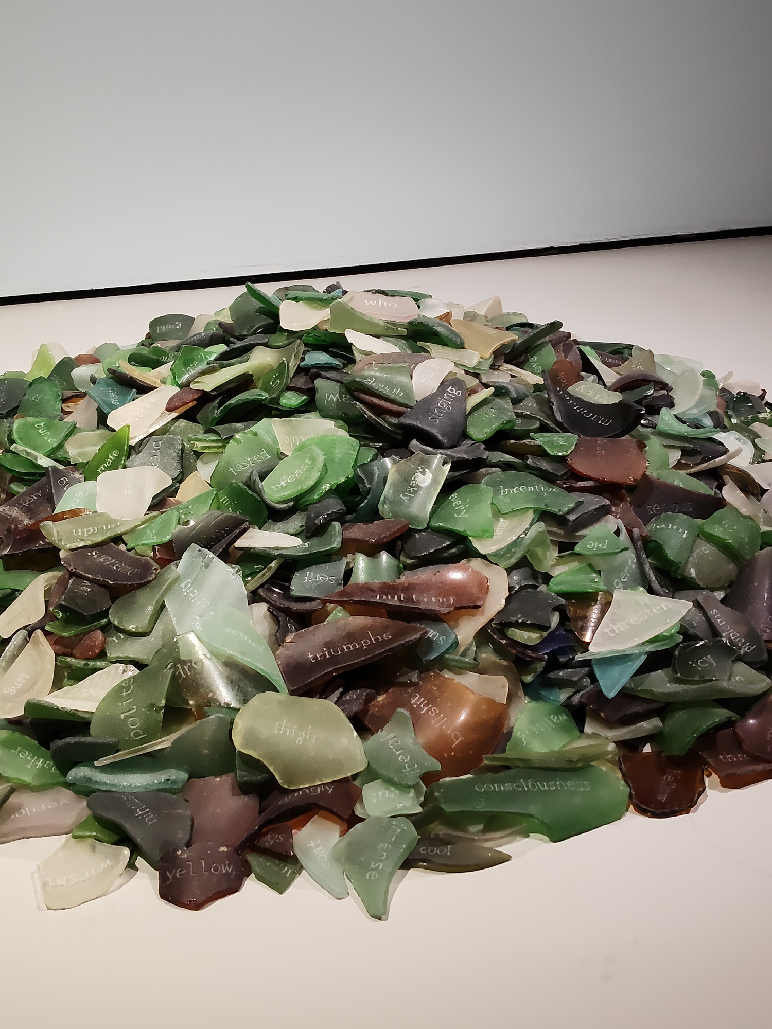 The image shows a pile of broken glass pieces of various colors including green, brown, and clear. Many of the glass shards have words etched onto them. The pile is on a white surface, with a white wall in the background. The composition suggests an art installation or display. No specific location in Singapore is identifiable from the image.