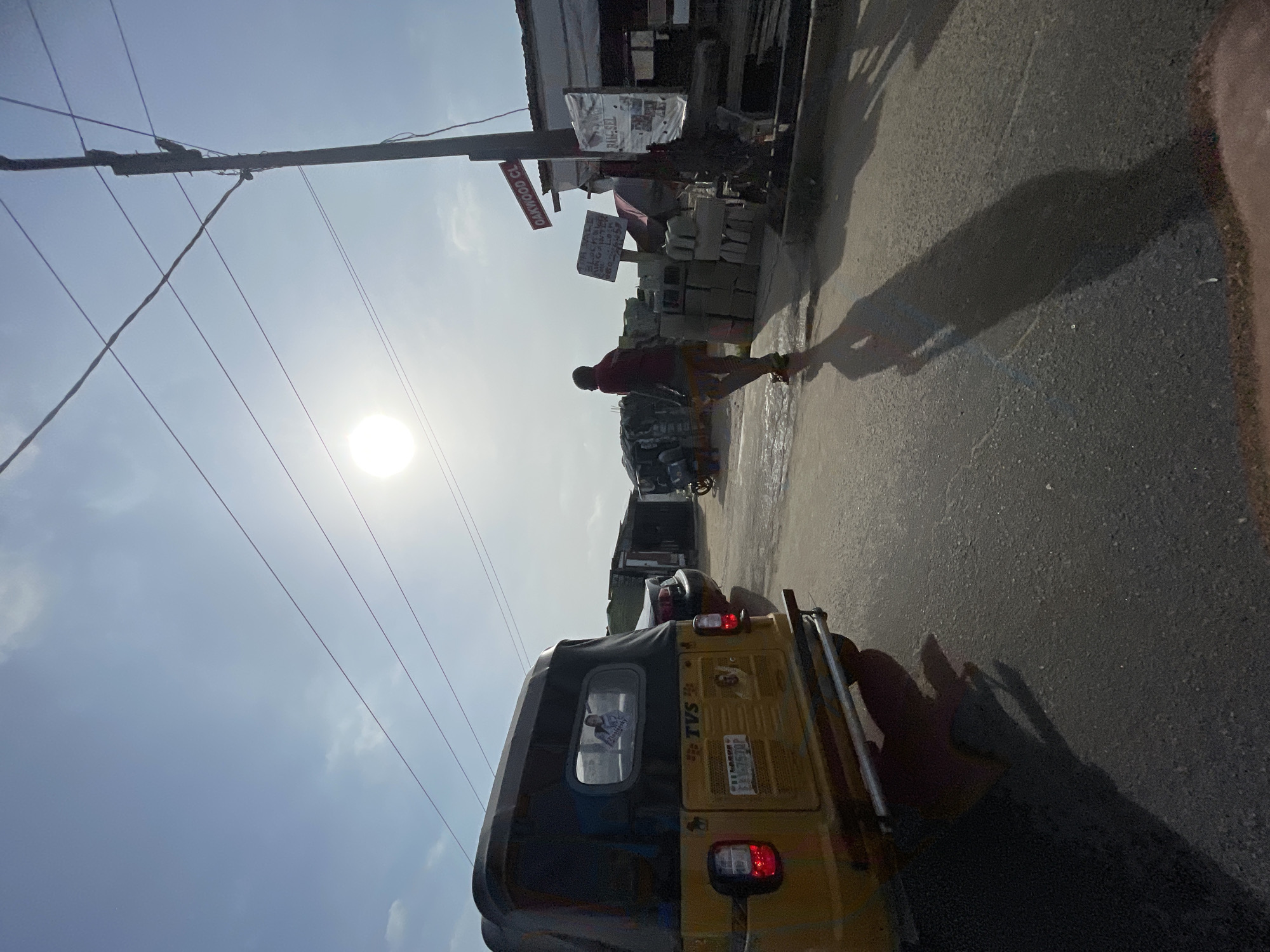 The image shows a street scene in Ado, Nigeria. A yellow "TVS" branded three-wheeled vehicle is the focal point, partially obscuring the view forward. A man in a red shirt and patterned shorts is walking along the side of the road, his shadow cast on the asphalt. A storefront to the side of the road advertises "OAKWOOD CL", and displays blocks and bags of materials. Overhead are power lines against a bright, sunny sky. Other vehicles can be seen further down the road.