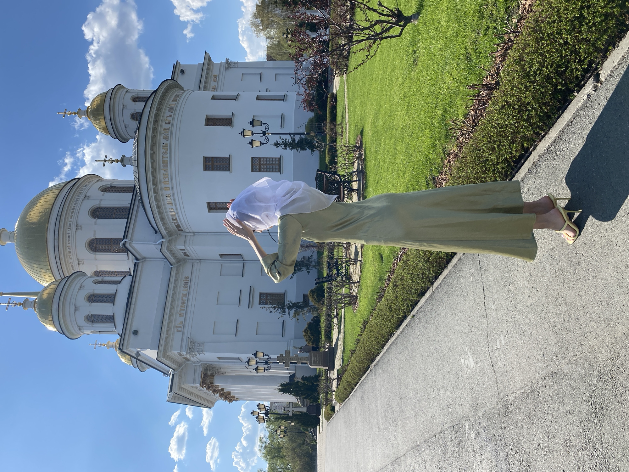The image shows a woman standing on a paved walkway near the Church on the Blood in Yekaterinburg, Russia. The woman is wearing a long, light green dress and yellow high-heeled sandals. She has a white headscarf covering her hair and is raising her hands towards her head.
The Church on the Blood, a large white building with golden domes, stands prominently to the left of the woman. The sky is blue with scattered clouds. Adjacent to the walkway, there is a well-maintained green lawn with shrubbery, trees, and benches.