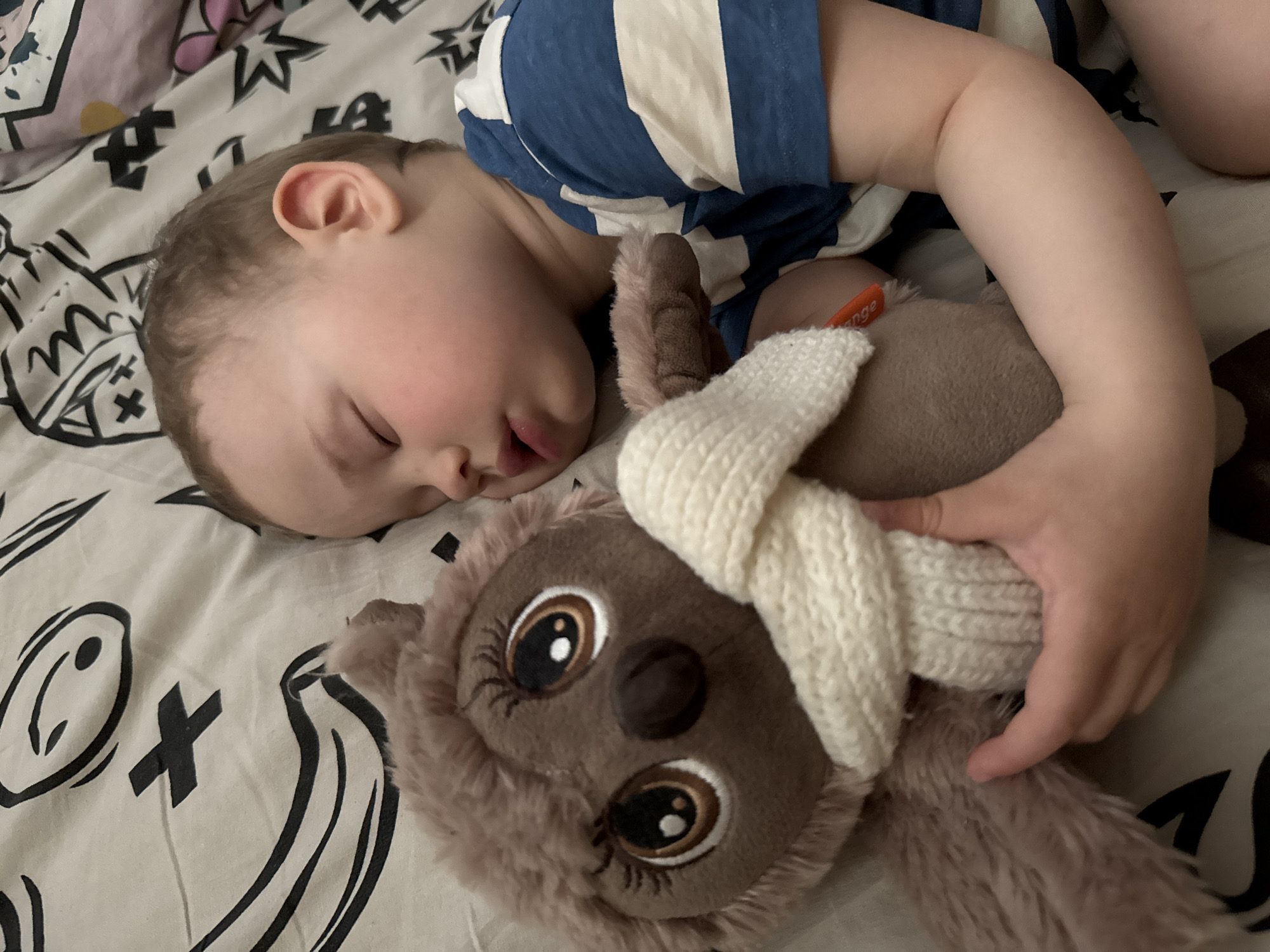 A toddler is asleep on a bed. The child is fair-skinned and wearing a blue and white striped shirt. The child is lying on their left side and clutching a brown stuffed owl with a white scarf around its neck. The bedspread is white with black graphic patterns. The location is Unknown, Unknown.