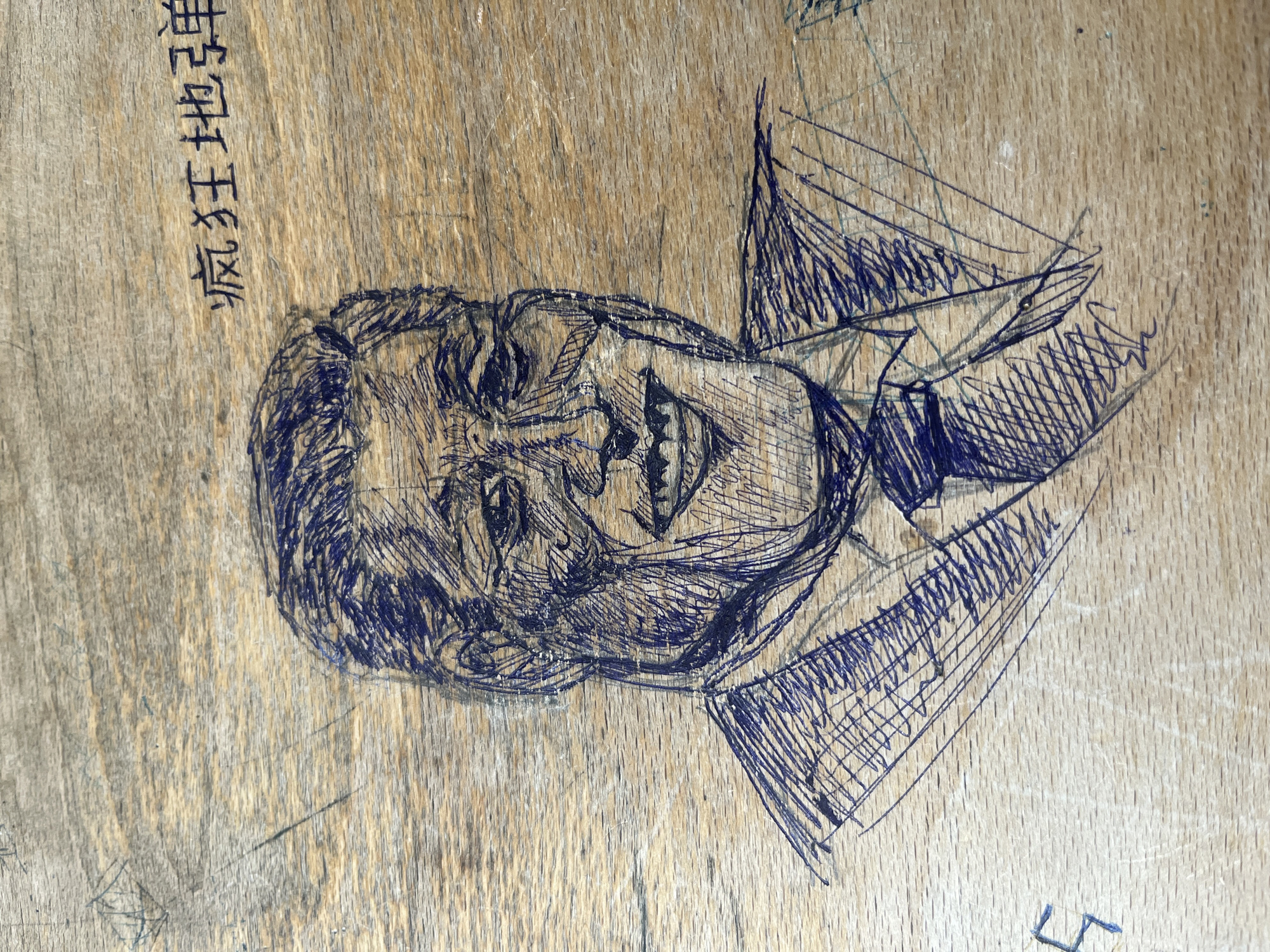 The image displays a close-up of a wooden surface, likely a desk or table, covered with pen markings and scratches. The dominant feature is a stylized drawing of a human face and upper torso, rendered in blue pen. The face appears male, depicted with an open mouth, revealing teeth, and an expressive, possibly laughing or shouting, countenance. Heavy hatching is used to give the sketch depth and define features like hair, wrinkles, and shading. The drawing includes parts of a collar and what might be a tie or suit lapels.
Above and to the left of the drawn head, a sequence of black characters, resembling East Asian script (likely Chinese), is visible, also marked onto the wood. The wooden surface itself is light-colored, showing a distinct grain pattern, along with numerous scratches, scuffs, and signs of wear, indicative of an aged or heavily used item of furniture. The artistic marks appear to be casual doodles or graffiti. The scene does not provide specific cues for time of day or weather, but the context suggests an indoor setting, possibly a classroom or similar environment in Sofia, Bulgaria.
