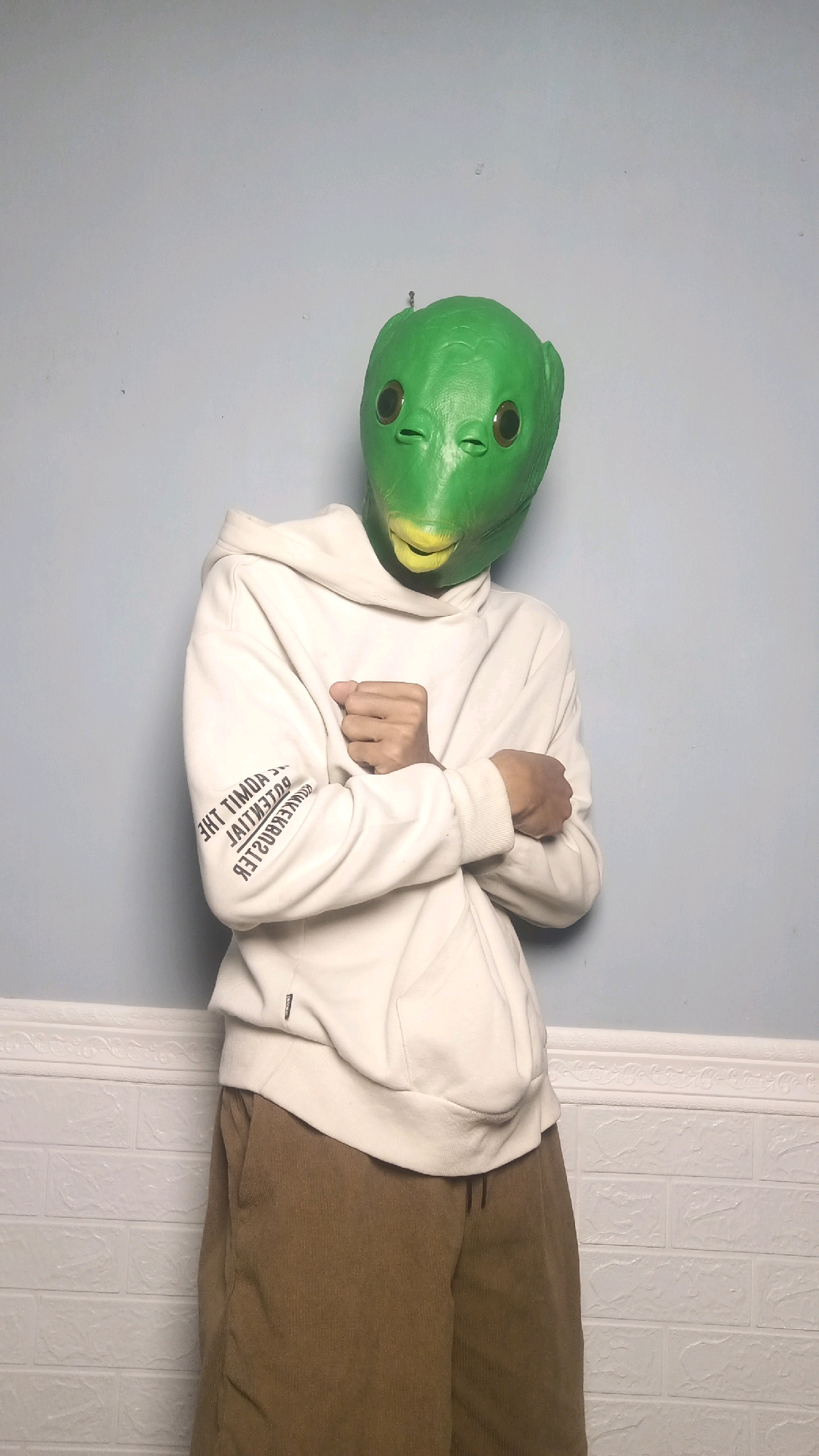 A person wearing a green alien mask, a white hoodie, and brown pants stands in front of a light blue wall. The hoodie has black text printed vertically on the left sleeve. The wall has a white textured border at the bottom. The person's arms are crossed in front of their chest. The setting is indoors and the lighting is even. There is no indication of the location being Kebumen, Indonesia.