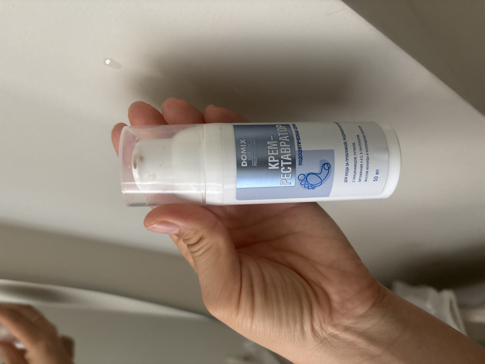A hand is holding a white plastic bottle of Domix Green Professional cream-restorer. The bottle has a blue label with text in Russian and a graphic of a foot. The label indicates the product is for problematic and irritated skin with niacinamide, rutin, vitamins A and E, monarda oil, and colloidal silver. The volume is 50 ml. The background is a plain, light-colored surface. The image appears to be taken indoors.