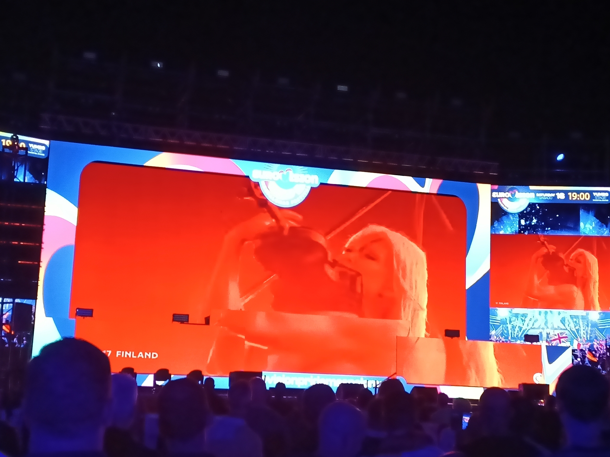 The image shows a large screen displaying the Eurovision Song Contest, as viewed by a crowd. The screen features the Eurovision logo, the name "Finland," and the number "17." The image on the screen appears to be a performance. A smaller screen shows a similar image along with a British flag and a crowd. The venue appears to be dark, and the lighting is predominantly blue. The location is most likely in San Bartolomé de Tirajana, Spain, based on the visual clues and the common staging locations for major events. The foreground is filled with the heads of audience members.