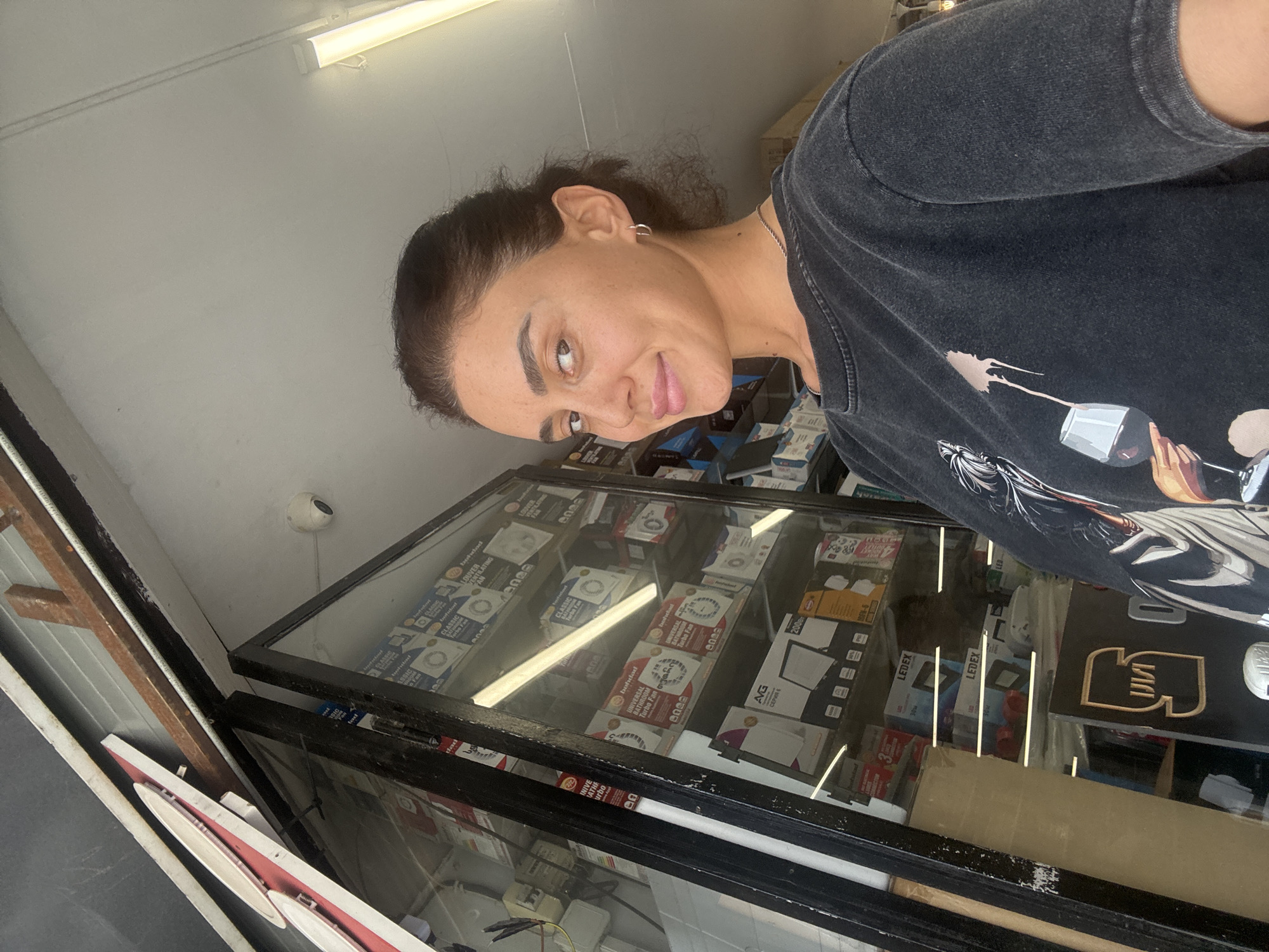 A woman with dark hair pulled back, wearing a dark graphic t-shirt and a silver necklace, stands in front of a shop window displaying various boxes of ventilation fans. She is looking towards the camera with a slight smile. The shop appears to be located in Tbilisi, Georgia, as indicated by the Cyrillic text visible on some of the items in the shop window and on a sign behind her. The shop's exterior features a dark metal frame around the window and a light-colored ceiling with fluorescent lighting. The overall scene suggests a casual, everyday moment captured outside a retail establishment.
