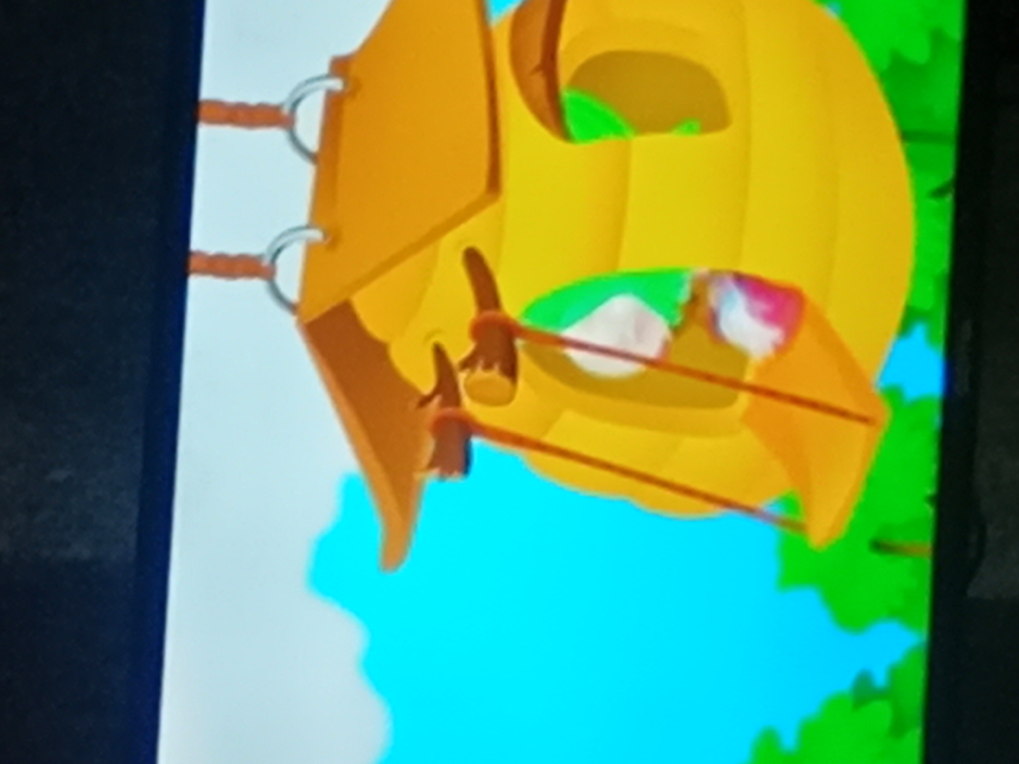 This media file is a photograph of a cartoon image displayed on a screen. The cartoon depicts a yellow, pumpkin-shaped structure that appears to be a hanging toy or ornament. Two orange ropes with metal rings are attached to the left side of the structure, suggesting it is suspended. Inside the structure, there are hints of green and white, possibly representing leaves or other decorative elements, and some blurry pink and white objects that could be flowers or candy. The background is a bright blue sky with some irregular white shapes, and a vibrant green leafy pattern is visible on the right side. The overall style is simple and colorful, typical of children's animation. There is no text visible in the image. The rotation of the image is unusual, with the subjects oriented sideways. The location context of Verkhniachka, Ukraine is not visually discernible from the image itself.
