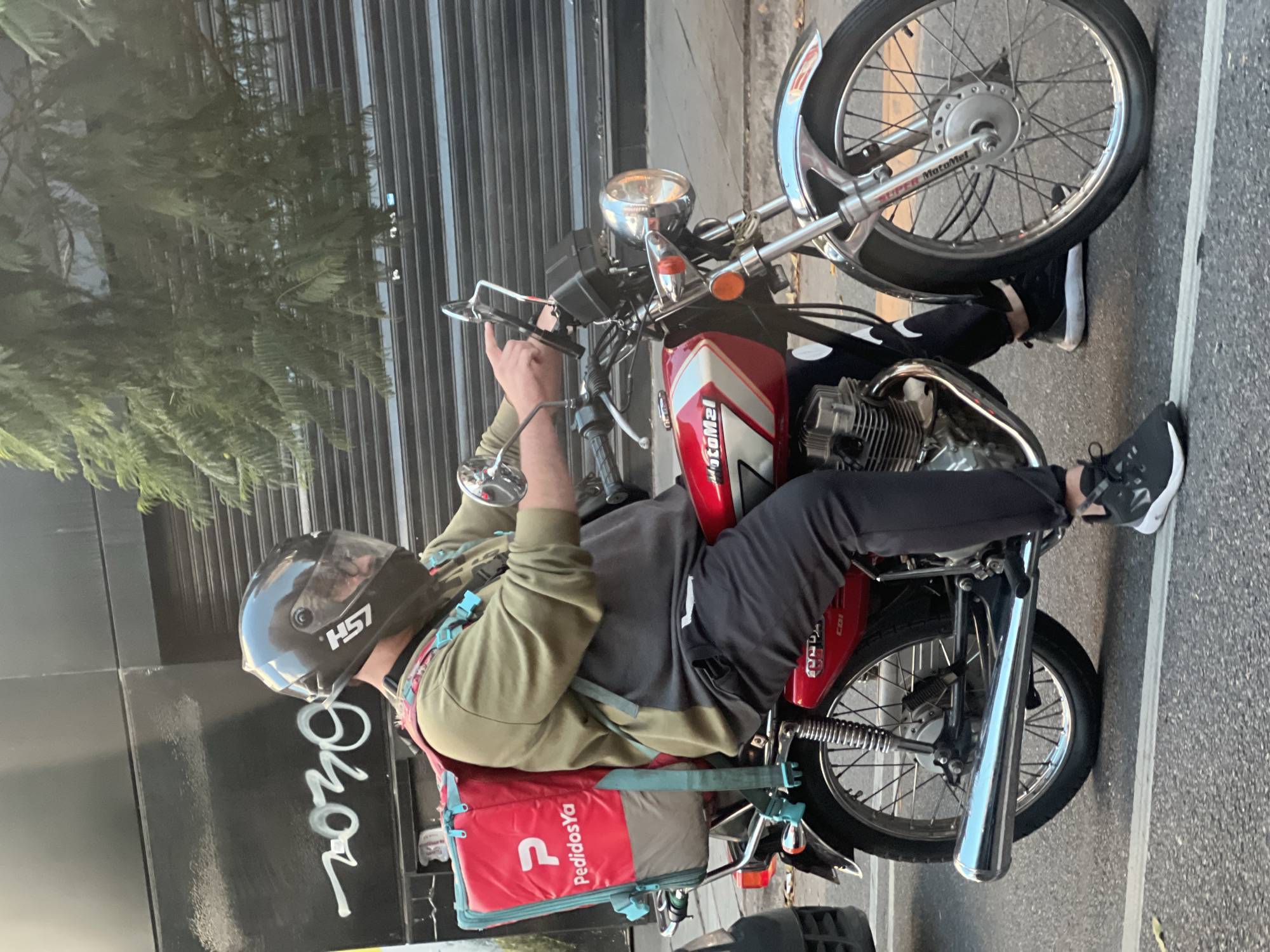 A delivery driver wearing a helmet, a gray sweatshirt with an olive green jacket, and black pants sits on a red motorcycle in what appears to be Buenos Aires, Argentina. He holds a phone in his left hand. The motorcycle is a "MotoMel" model. Behind him is a red delivery bag labeled "PedidosYa." The backdrop includes a building with graffiti, a rolled-up shutter, and a tree.