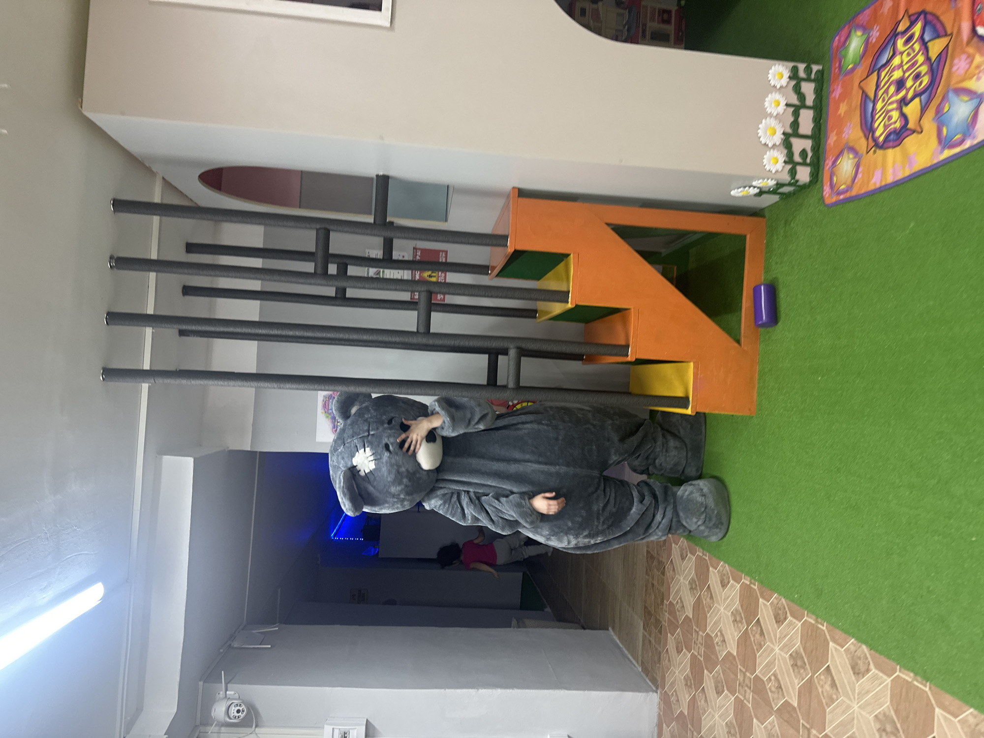 An indoor scene captured from a slightly elevated perspective shows a playful environment, possibly a children's entertainment center. The image is rotated 90 degrees clockwise.
In the foreground and center, a person wearing a full-body, grey teddy bear costume stands. The costume features a white patch on the teddy bear's head and its hands are raised, covering the face, suggesting shyness or playful hiding. The costume's feet are large and plush.
To the right of the teddy bear is a multi-level orange and green play structure resembling steps or platforms, with several black vertical bars acting as a barrier or climbing apparatus. The floor is divided into two distinct sections: a section of green artificial turf on the right and a patterned brown and tan tiled floor on the left. On the green turf, near the play structure, is a small purple cylindrical object. Further to the right on the turf, a colorful rectangular mat with "Dance Magic" or a similar phrase, surrounded by stars and abstract shapes, is visible. Behind the play structure, white fake daisy decorations line a ledge.
In the background, a dark entryway or hallway is visible. Another person, possibly a child or small adult, wearing a pink top and light-colored pants, is seen walking away into this darker area. Above the hallway entrance, a bright fluorescent light illuminates the ceiling, where a white security camera is also mounted.
Visible text includes "Dance Magic" on the mat and partial text on a sign behind the black bars, which appears to be a rules sign with Cyrillic words like "ПРАВИЛА" (RULES) and "ОБЯЗАТЕЛЬНО" (MANDATORY).