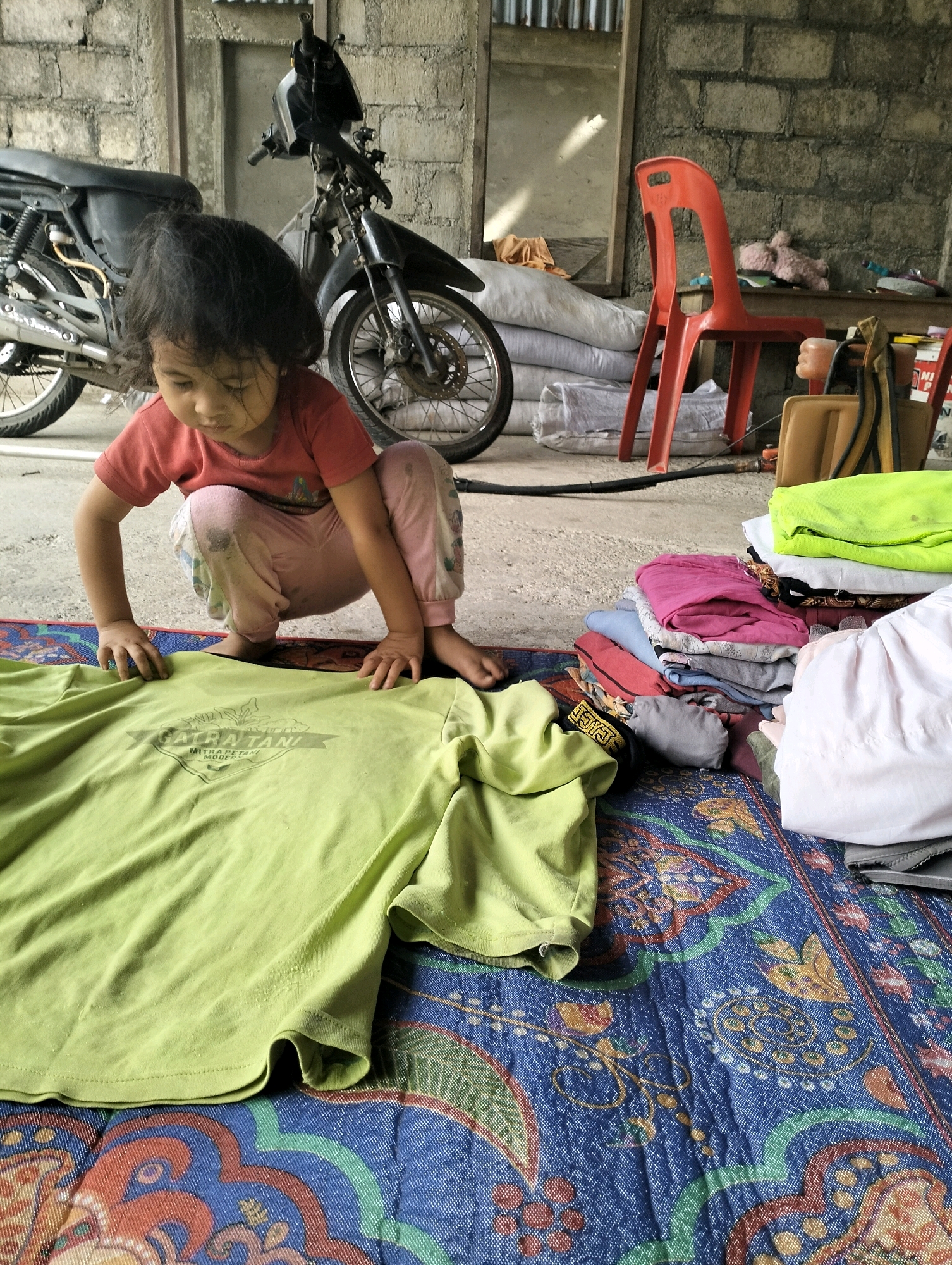 A young girl, likely Indonesian, is crouched on a colorful woven mat. She is wearing a pink t-shirt and light-colored pants. Her hands are resting on a large, lime green t-shirt spread out in front of her. The t-shirt has a dark logo with text that is difficult to read clearly but appears to be in Indonesian. In the background, a motorcycle is parked against a weathered brick wall. Piles of folded clothes are stacked to the right of the girl, and a red plastic chair is visible further back. The environment suggests an outdoor or semi-outdoor space, possibly a courtyard or a simple dwelling, with a concrete floor. The overall scene depicts a domestic moment, with the child interacting with laundry in a humble setting in Pekan Hilisimaetano, Indonesia.