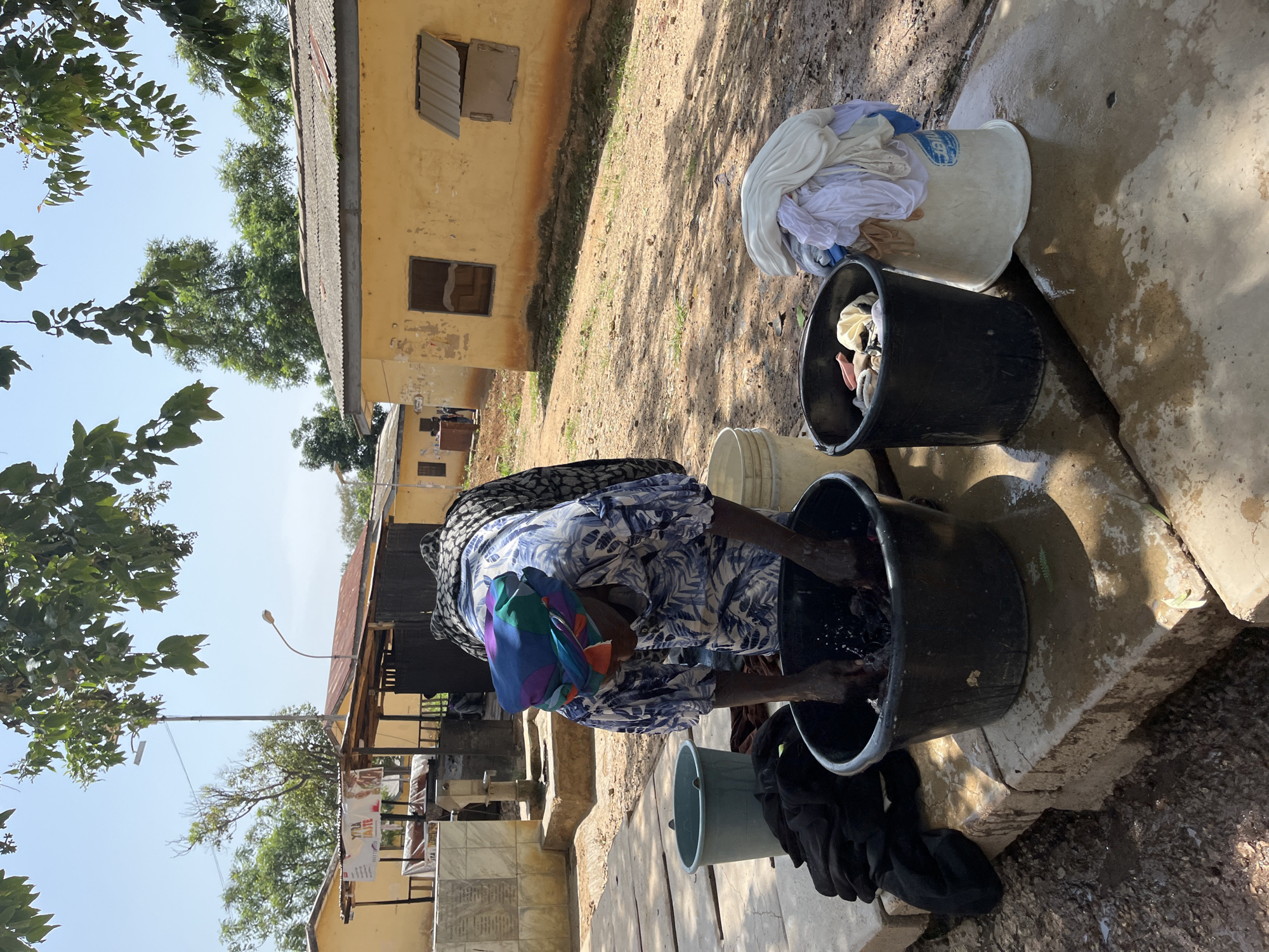 A person is washing clothes in large buckets outdoors. The scene appears to be in a village setting, with a weathered yellow building in the background that has barred windows. The ground is dirt and concrete, and there are trees with green leaves visible. Several buckets are present, some filled with laundry, one containing water. The person is wearing a blue and white patterned dress and a colorful headwrap. The sunlight suggests it is daytime. The activity is likely a common household chore. There is no visible text in the image.