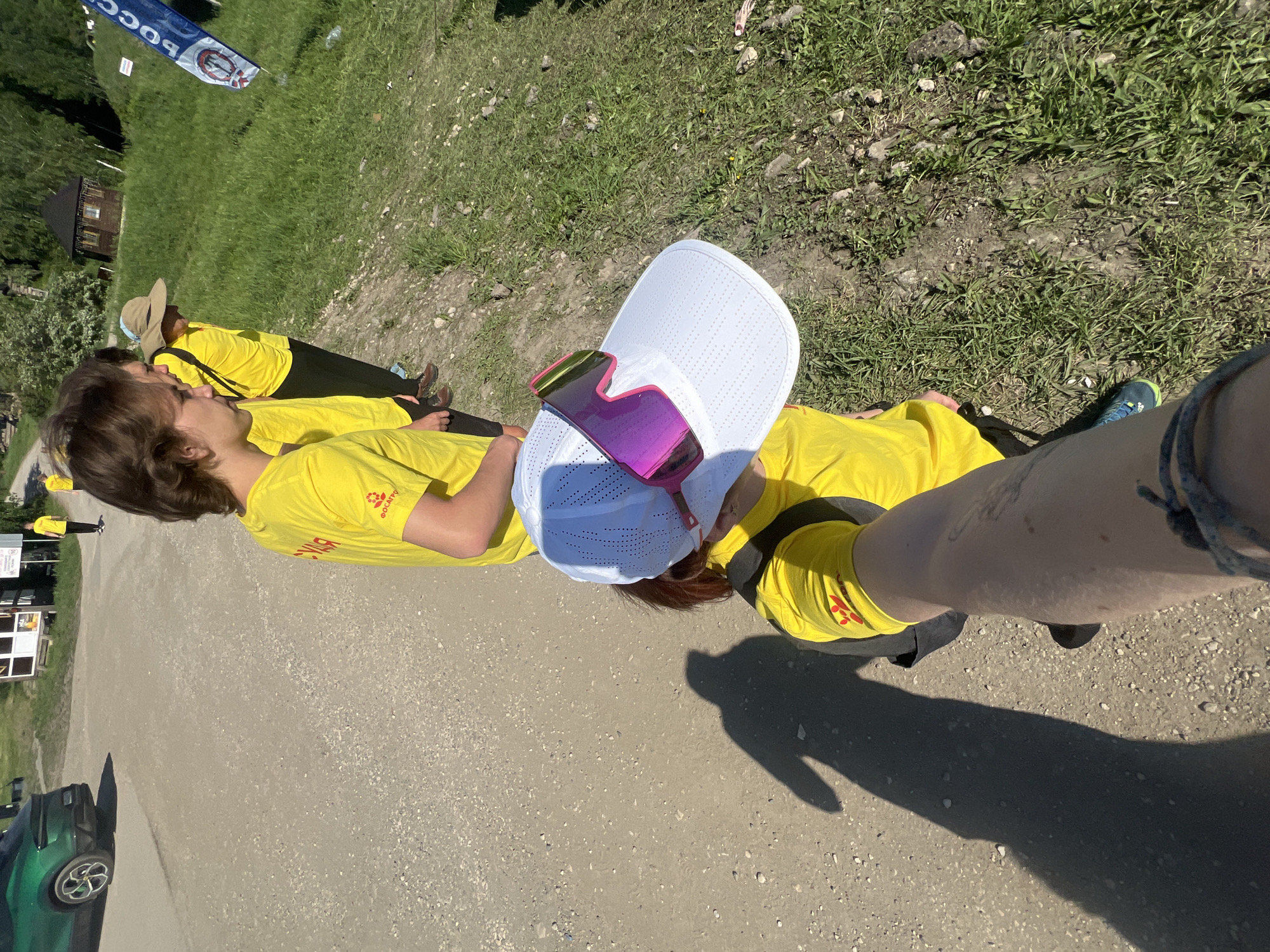 A low-angle medium shot shows several people wearing bright yellow shirts standing in a line outdoors. The shirts appear to have Russian writing on them, and at least one person is wearing a white visor with pink-tinted sunglasses on top. The person closest to the camera is taking a selfie. They're standing on a dirt or gravel road with grass and trees in the background. A building is visible in the background, and a flag with Russian writing is nearby. Based on the writing on the shirts and the flag, the location is possibly in Khvalynsk, Russia.