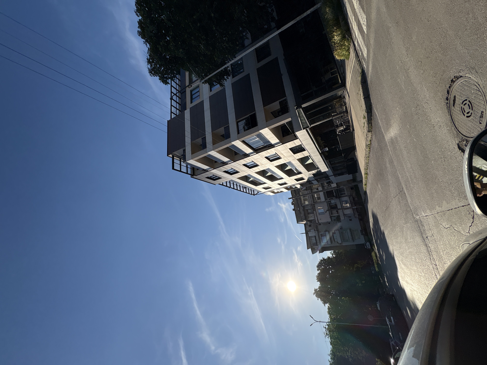 The image depicts a street scene in Vratsa, Bulgaria, under a clear blue sky with scattered cirrus clouds. The sun is visible, casting long shadows and creating a lens flare. In the foreground, the curve of a car's hood and a side mirror are visible, suggesting the photo was taken from inside a vehicle. The street is asphalt with white pedestrian crossing markings. A manhole cover is centered in the lower right portion of the frame.
Dominating the right side of the image is a modern, multi-story apartment building with a sleek design featuring dark horizontal bands and balconies. To the left of this building, an older, taller apartment building with a more traditional, weathered facade stands. Dense green trees line the street, obscuring parts of the older building and casting shadows on the road. In the distance, power lines stretch across the sky. The overall impression is of an urban environment bathed in the warm light of late afternoon or early morning.