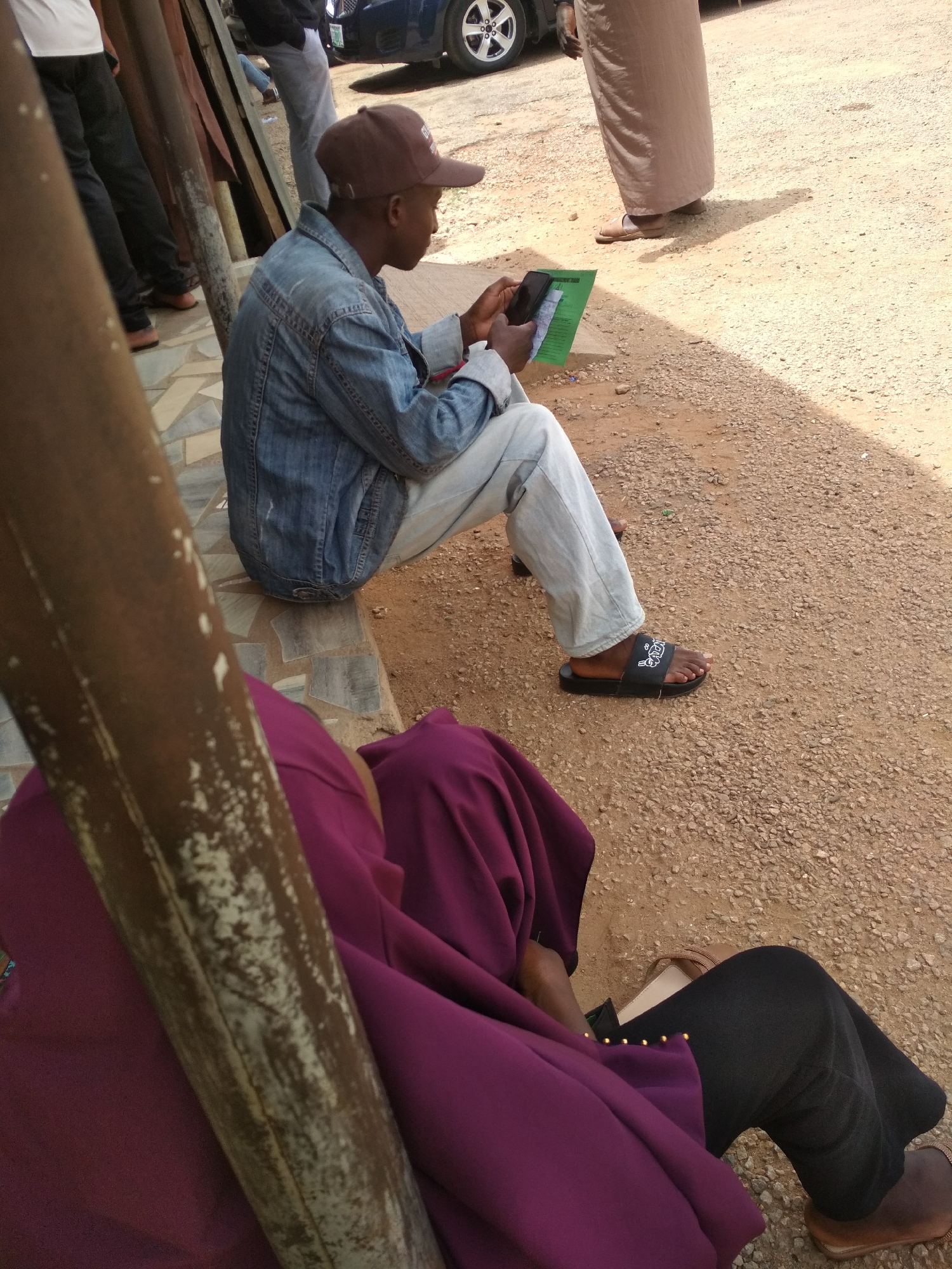 In Gura Top, Nigeria, the image captures a moment outside a building. A man wearing a brown baseball cap, denim jacket, and light-colored pants sits looking at his phone while holding a green paper document. He is wearing black sandals. Nearby, a person draped in a purple garment sits with black pants and brown sandals. Other individuals are also present in the background. The ground is gravel.
