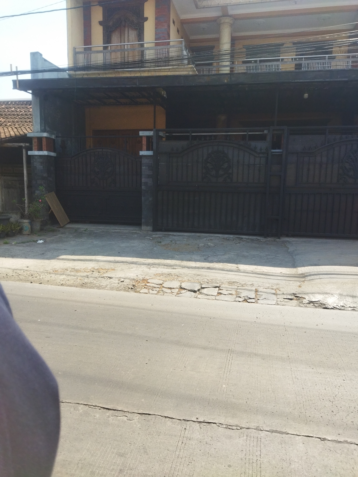 The image shows a wide shot of a house in Juwiring, Indonesia, with a large black metal gate in front. The house has multiple stories, with balconies and intricate architectural details on the upper floors. The gate is ornate, featuring a circular design with what appears to be a tree or floral motif. To the left of the gate, there are several potted plants and a wooden plank leaning against the gatepost. The ground in front of the gate is a paved driveway and sidewalk, with some cracking and disrepair visible. A street runs in front of the property, with a portion of a person's leg visible in the lower left corner of the frame, suggesting a street-level perspective. The overall scene depicts a residential property in a tropical or sub-tropical climate, given the sunlight and vegetation.