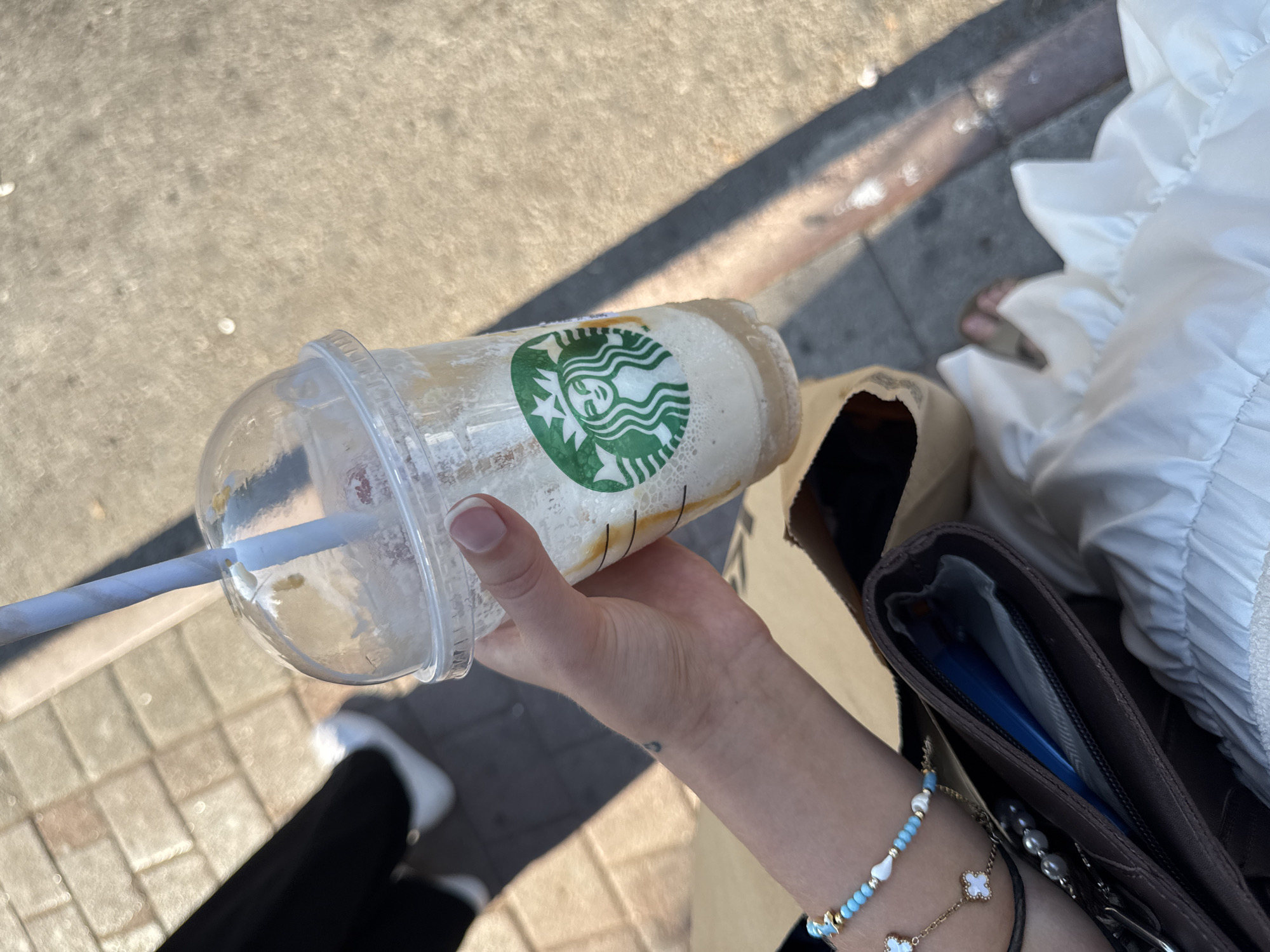 The image shows a person holding a partially consumed Starbucks iced beverage in a clear plastic cup with a domed lid and a white paper straw. The Starbucks logo is visible on the cup. The person is wearing bracelets on their wrist, one of which is light blue with white beads and charms. A brown bag is also partially visible, containing some items. The background consists of a sunlit sidewalk with cobblestone patterns and shadows. The person is wearing black pants and white sneakers. The context of Antalya, Turkey, is not evident from the image itself.