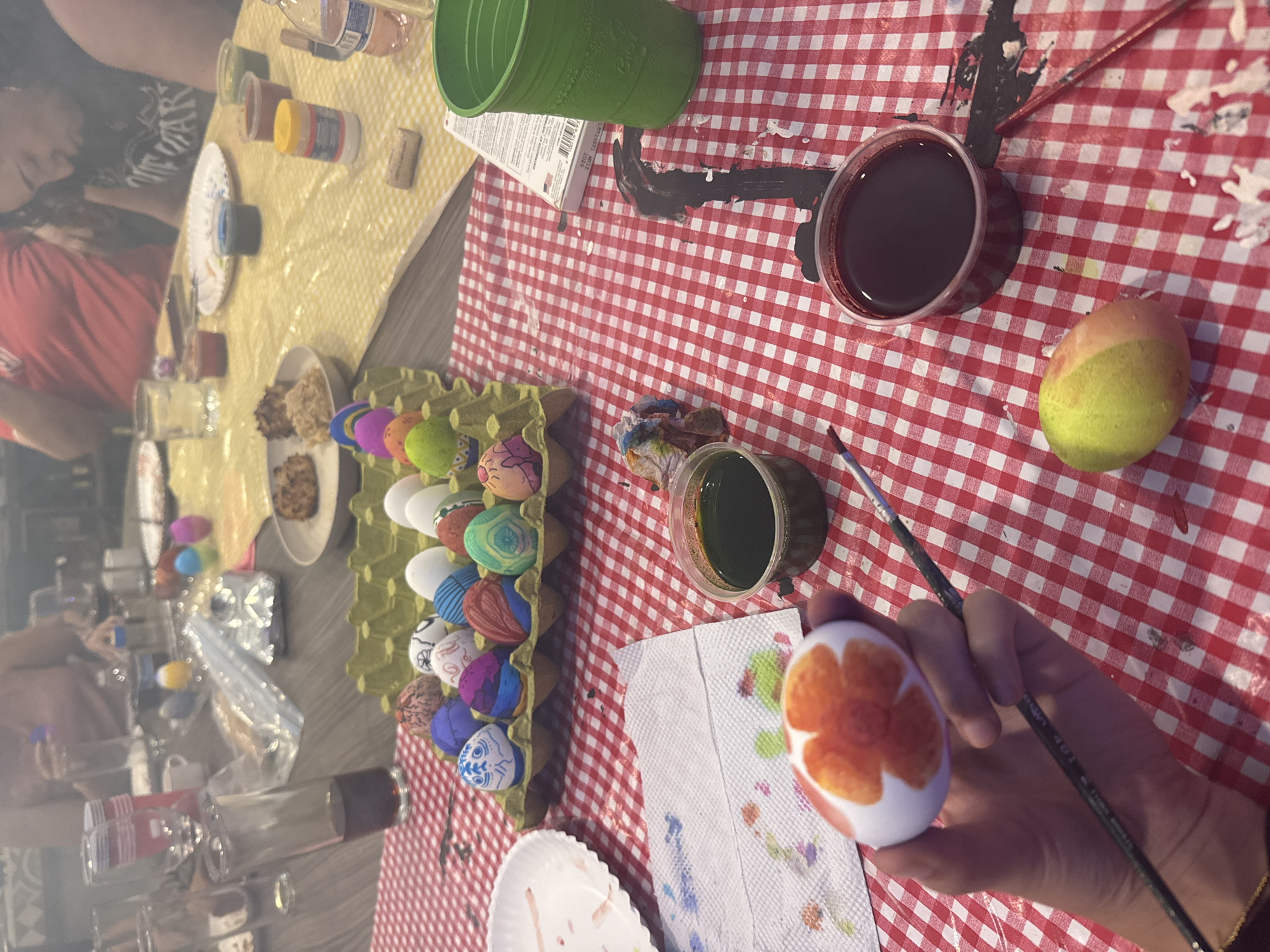 The image shows a person painting an egg. The egg is white with an orange flower design. The person is holding the egg in their left hand and a paintbrush in their right hand. There are other painted eggs in a carton next to the person. There are also cups of paint and other supplies on the table. The table is covered with a red and white checkered tablecloth. The location is likely a home or art studio in San Bernardino Tlaxcalancingo, Mexico, based on the presence of traditional Mexican crafts like painted Easter eggs.