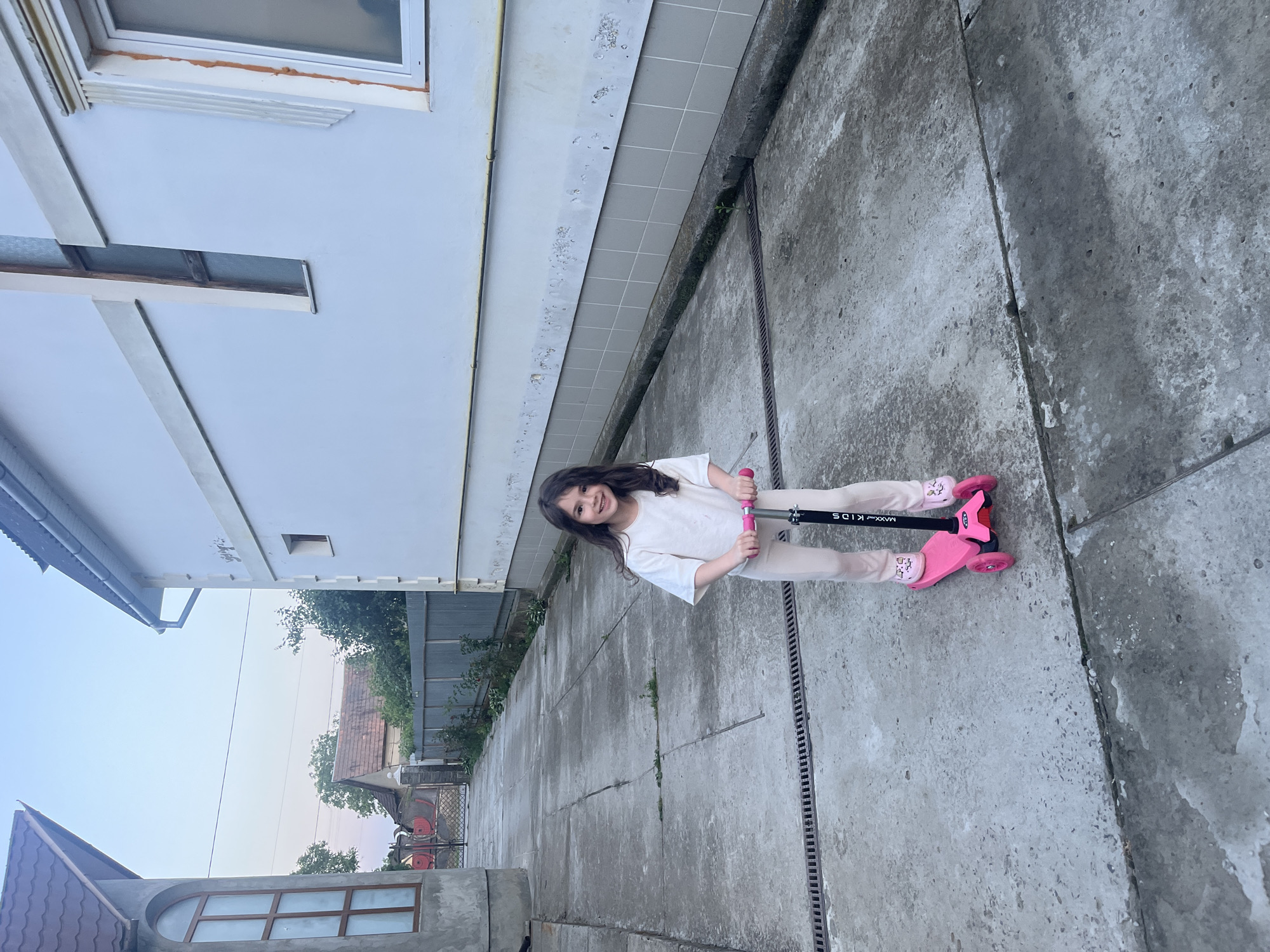 A young girl with long, dark hair is depicted standing on a pink scooter on a concrete path in Tarnivtsi, Ukraine. She is wearing a white t-shirt and light-colored pants, with patterned socks visible above her bright pink footwear. The girl is smiling broadly at the viewer, holding the black handlebars of her scooter, which has "MAXX KIDS" printed on its stem.
The setting is an outdoor area, likely a residential yard or walkway. To the girl's left, a white building with visible windows and a tiled section along its base runs parallel to the concrete path. A metal drainage grate is embedded into the concrete surface to her right. In the background, beyond a fence, other buildings with brown roofs and lush green trees are visible under a bright, clear sky, indicating daytime, possibly late afternoon or early evening. The concrete path shows signs of weathering and use. The overall scene captures a moment of childhood joy against a backdrop of a typical residential environment.