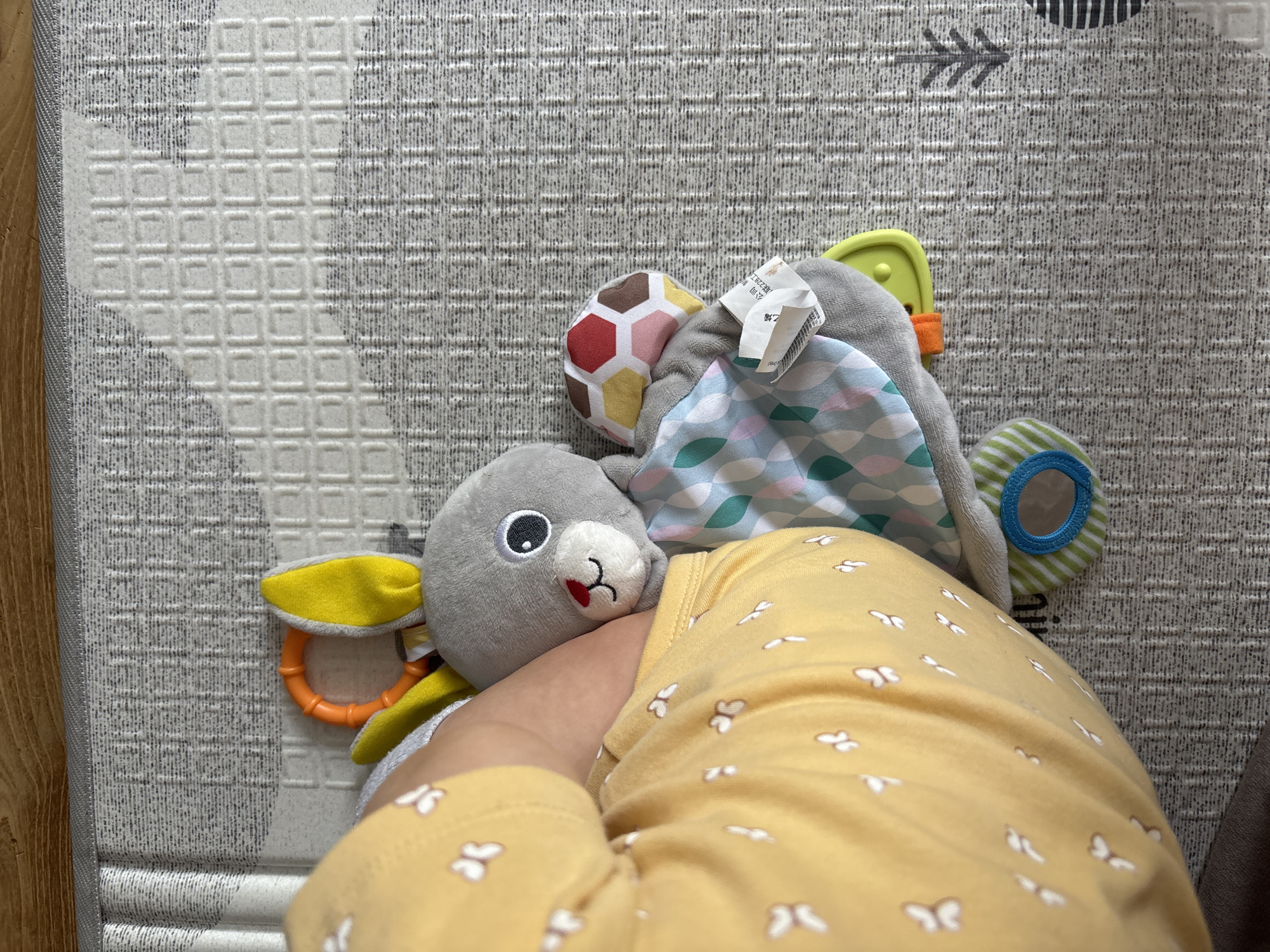 The image displays an overhead view of an infant's arm and partial torso, dressed in a yellow long-sleeved garment patterned with small white butterflies. The arm is positioned on a grey and white playmat with various textured geometric patterns, including squares and horizontal lines. A section of a wooden floor is visible along the top edge of the frame.
The infant's arm is interacting with or resting near a multi-sensory plush bunny toy. The bunny is primarily grey with bright yellow ears, a red nose, and prominent black and white eyes. Attached to the toy are several features: an orange plastic teether ring, a soft fabric section patterned with teal, green, and pink ovoid shapes, another fabric section composed of multi-colored hexagons (red, yellow, brown, pink), a white manufacturing tag featuring text in an Asian script (likely Chinese), and a striped green and white fabric ring containing a small circular mirror.
The overall scene is an indoor domestic environment. No specific architectural features, landmarks, or identifying elements are present in the image to corroborate a location in Sofia, Bulgaria.