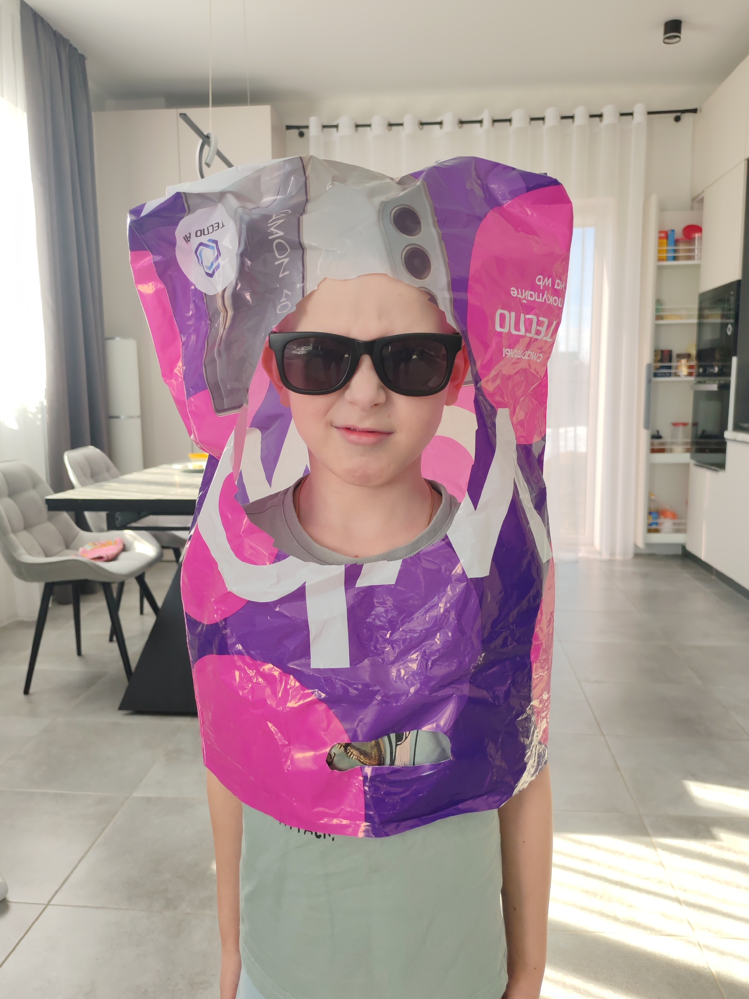 An interior scene depicts a young individual, appearing to be a child, standing centrally in what is identified as a modern residential space in Chaikovsky, Russia. The child faces the camera directly, wearing dark sunglasses and a light green t-shirt. They are partially encased in a crumpled, oversized plastic bag, predominantly purple and pink, with white accents. The bag features prominent "TECNO" branding and inverted Cyrillic text, appearing to read "SMARTPHONES" and "AMON" (likely part of "CAMON"). A cutout on the top of the bag is positioned over the child's head, simulating the appearance of a multi-lens camera array found on mobile phones.
The background reveals an open-concept living area with light gray tiled flooring and white walls. To the left, a dining area is visible, featuring a rectangular table with a dark surface and black legs. Two light gray upholstered chairs with black legs are present, one of which holds a pink object, possibly a small bag or toy. Further back, a light-colored appliance or cabinet is partially visible, alongside gray curtains covering a window. To the right, a contemporary kitchen area features white cabinets, an integrated appliance (possibly an oven), and an open shelving unit displaying various colorful household items. A large window with sheer white curtains on a black rod allows bright natural light to enter, casting distinct shadow stripes on the floor. A black track light fixture is mounted on the ceiling, and a pendant light fixture with black rings hangs above the dining table.