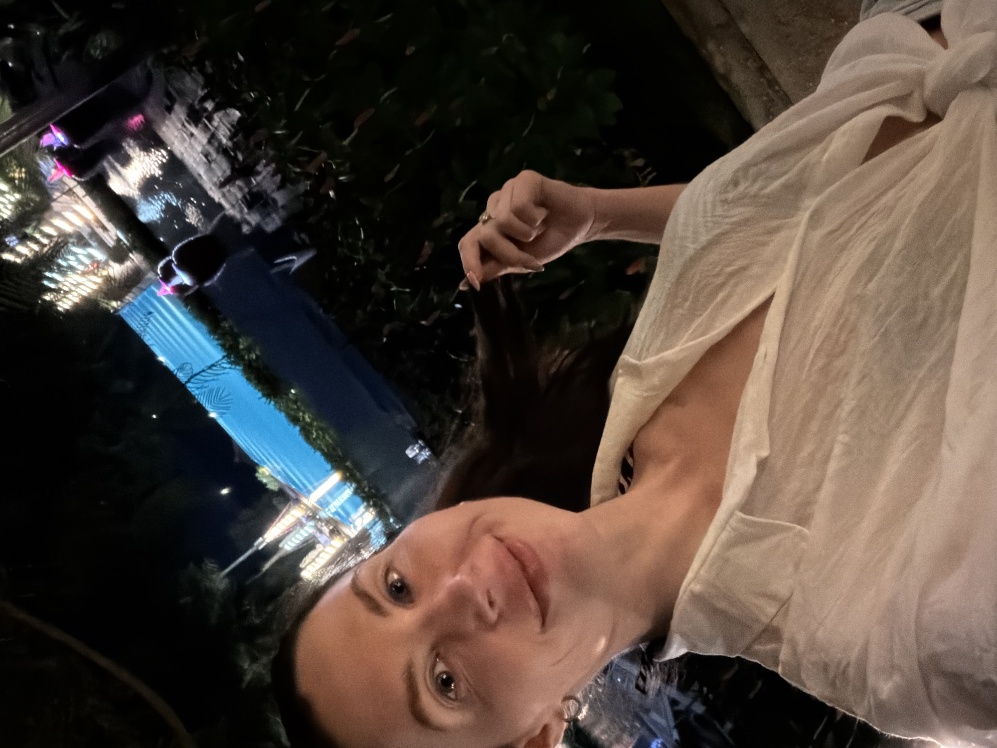 The image depicts a close-up, waist-up selfie of a woman taken at night in an outdoor setting in Dong Ninh Hoa Ward, Vietnam.
The woman, approximately in her 30s, occupies the left foreground, looking directly at the camera with a gentle smile. She has long, dark brown hair, parted on her left, with some strands resting on her right shoulder. Her skin is fair, and a small hoop earring is visible in her left ear. She is wearing a loose-fitting, light cream or off-white collared shirt, unbuttoned to reveal a glimpse of a dark garment underneath. Her right hand is subtly raised, with fingers curled, appearing to touch or hold a strand of her hair near her ear. A gold-toned ring with a central stone is present on her right ring finger.
The background reveals an illuminated outdoor leisure area. A prominent bright blue swimming pool or water feature spans the mid-ground horizontally, accented by internal blue lighting along its edge. Lush, dark green tropical foliage and palm tree silhouettes frame the water feature against the dark night sky. To the left of the water, several structures with multiple warm-toned string lights or spotlights are visible. To the right, a dark, low barrier or ledge separates the foreground from the illuminated area. Beyond the blue water, reflections of light are visible on what appears to be another small body of water or wet surface. Two indistinct, dark human figures are present near the far side of the blue water feature, one standing upright, the other further back. Scattered small pink and purple glow lights are positioned on the ground or low structures in the right background. The overall scene suggests a resort, hotel, or upscale restaurant environment during the evening.