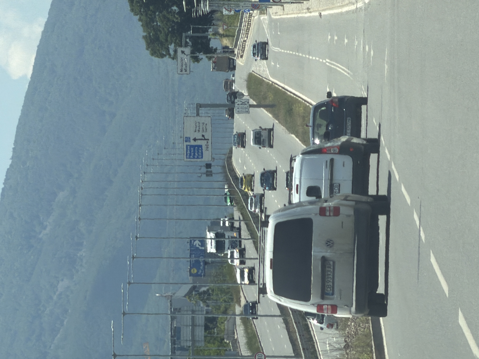 The image depicts a multi-lane highway leading into a city, likely Sofia, Bulgaria, given the visible signage and European-style vehicles. The highway is flanked by grassy embankments and modern street lighting. In the foreground, a white Volkswagen Caddy van with license plate "CB 9393 AP" is prominently visible, followed by another white van. Several other vehicles, including cars and a truck, are on the highway, some in motion and others stationary. The road is marked with white lane dividers. In the background, a large mountain range covered in vegetation dominates the skyline, with a partly cloudy sky above. A street sign indicating distances to "Pernik," "Kyustendil," and "290M" is visible on the left, further suggesting a location in Bulgaria. Another sign on the right has text in Cyrillic, with "MADARA" and "MEADE" visible. The overall scene is one of typical urban highway traffic under daylight conditions.