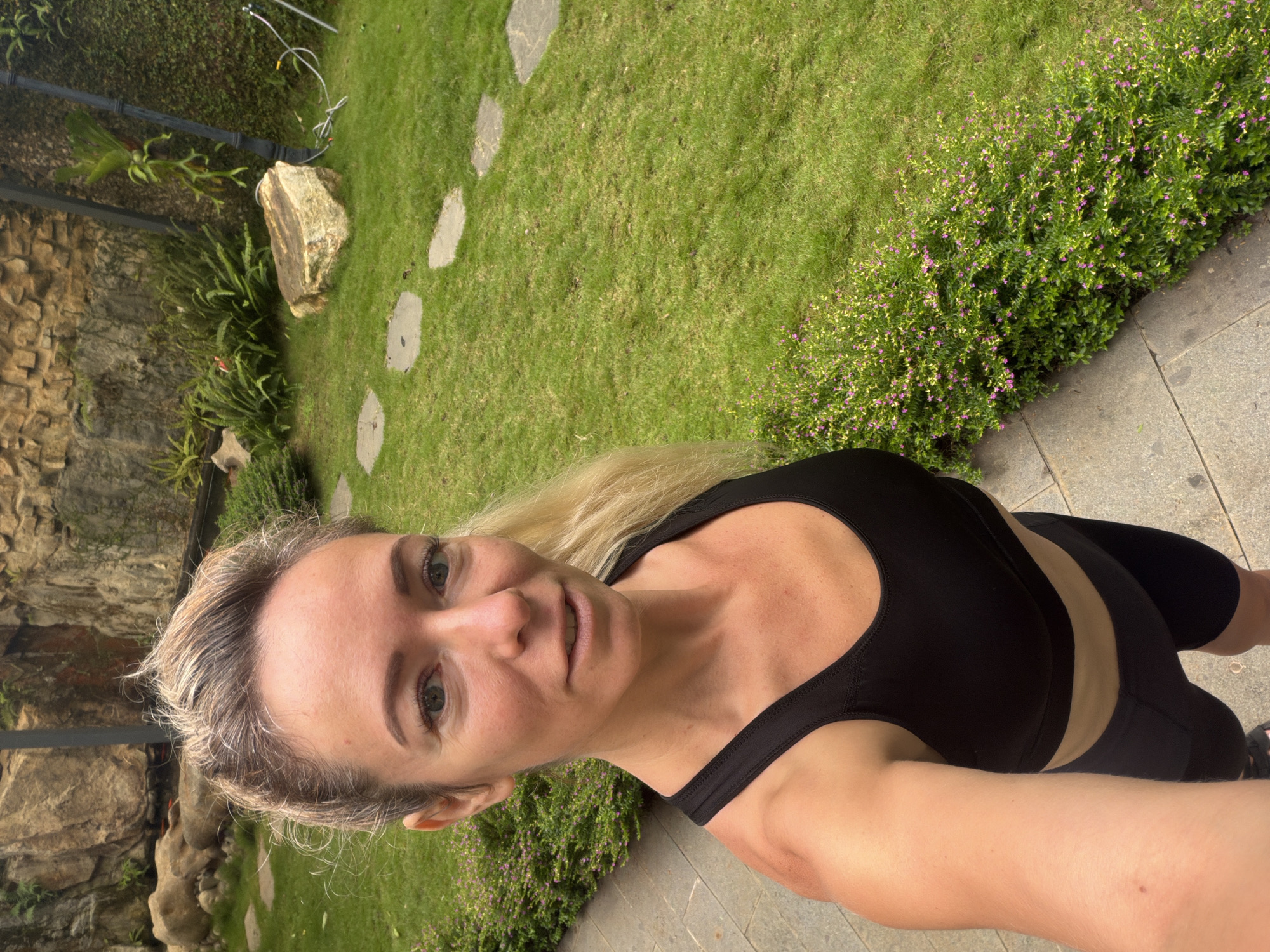 A woman with blonde hair pulled back into a ponytail and wearing a black sports bra and black shorts is taking a selfie in a garden. She has fair skin and appears to be outdoors. The background shows a stone wall, a lush green lawn, and some flowering bushes. In the distance, there are large rocks and a lamp post with some plants growing on it. The ground in the foreground is paved with stone tiles. The image was taken in the city of Xuân Trường - Đà Lạt, Vietnam.