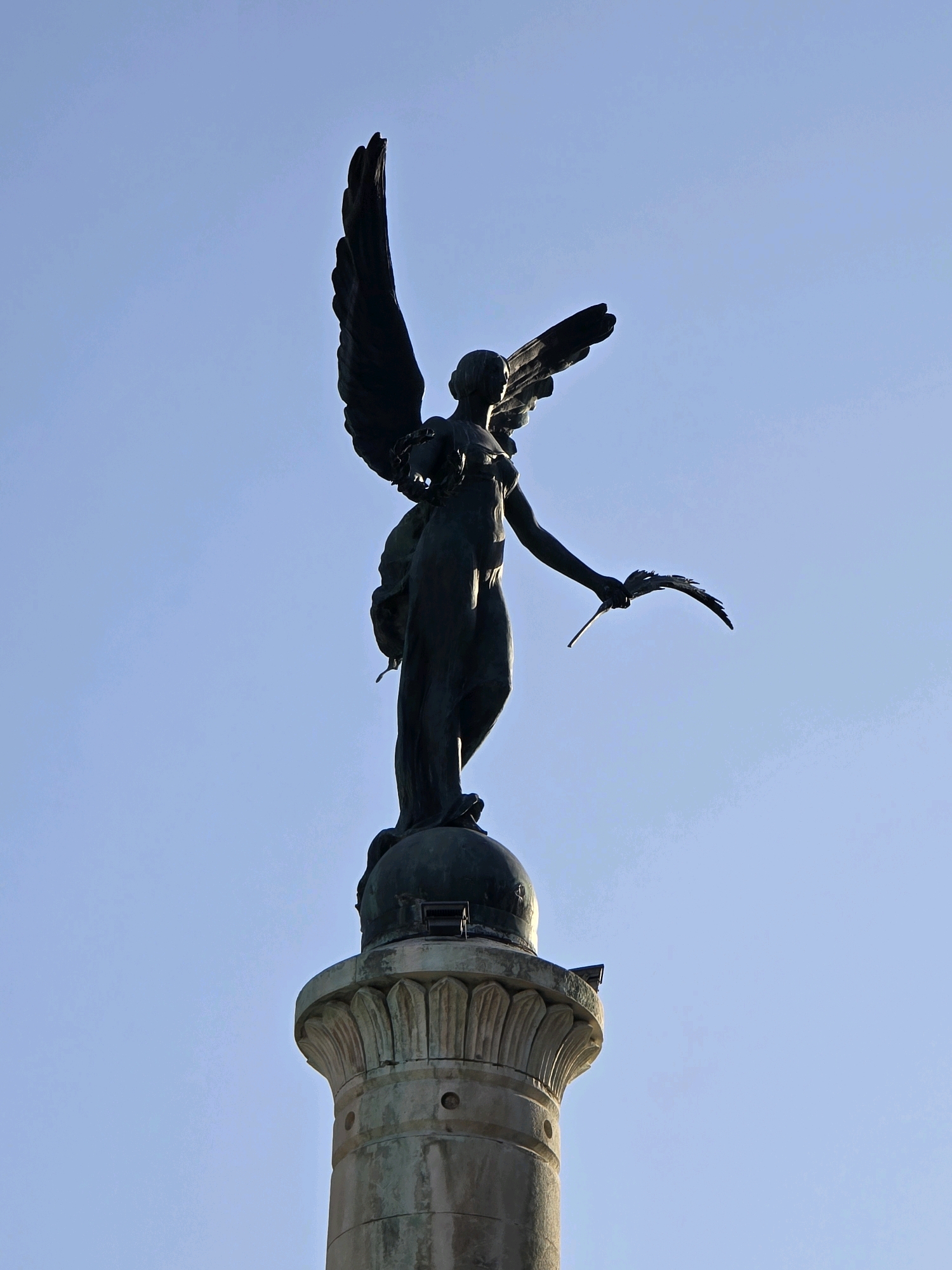 The image shows a bronze statue of a winged female figure, likely a representation of Victory or an angel, standing atop a fluted column. The statue is dark in color and appears to be made of weathered bronze. The figure is depicted with its wings spread, and in its right hand, it holds a laurel wreath. The statue stands against a clear, light blue sky. The column is made of stone, with decorative carvings at its top. The overall scene suggests an outdoor monument. This monument is located in the Plaza de las Batallas in Jaén, Spain.