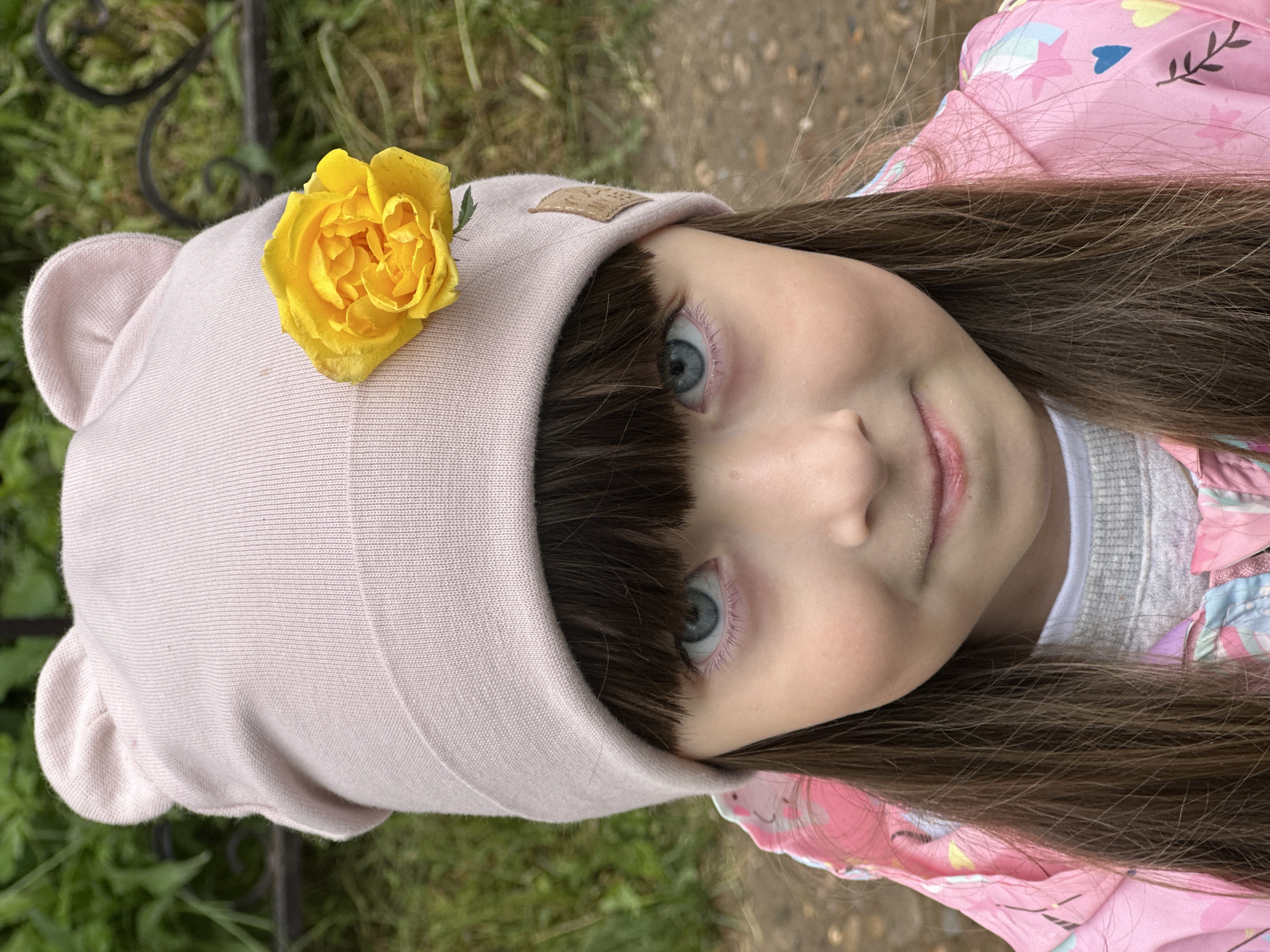 A young girl with blue eyes and dark brown bangs looks directly at the camera, exhibiting a slight, closed-mouth smile. She wears a light pink knit beanie with two subtle, rounded protrusions resembling bear ears on top. A vibrant yellow rose is affixed to the right side of the beanie (viewer's left). Her long, dark brown hair falls over her shoulders. She is dressed in a pink jacket adorned with multi-colored patterns, layered over a white shirt and a grey knit sweater, both partially visible. The background is softly out of focus, revealing green foliage to the upper left and a brown, earthen ground or path to the right. A dark, curvilinear metal object, possibly part of a fence, is faintly discernible in the upper right background. Natural light illuminates the scene evenly.