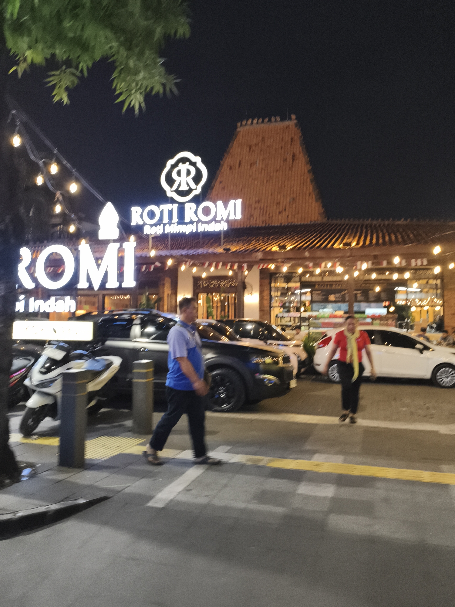 The image captures a nighttime scene in front of a "Roti Romi" establishment, identified as being in South Jakarta, Indonesia. The building features a prominent wooden roof structure with multiple levels and is adorned with numerous string lights. In the foreground, a sidewalk area shows a man in a blue and white shirt walking to the left, and a woman in a red shirt and yellow scarf walking to the right. Several vehicles, including a white scooter, a dark-colored SUV, and a white sedan, are parked or present in the vicinity. The overall atmosphere suggests a busy, possibly commercial area at night.