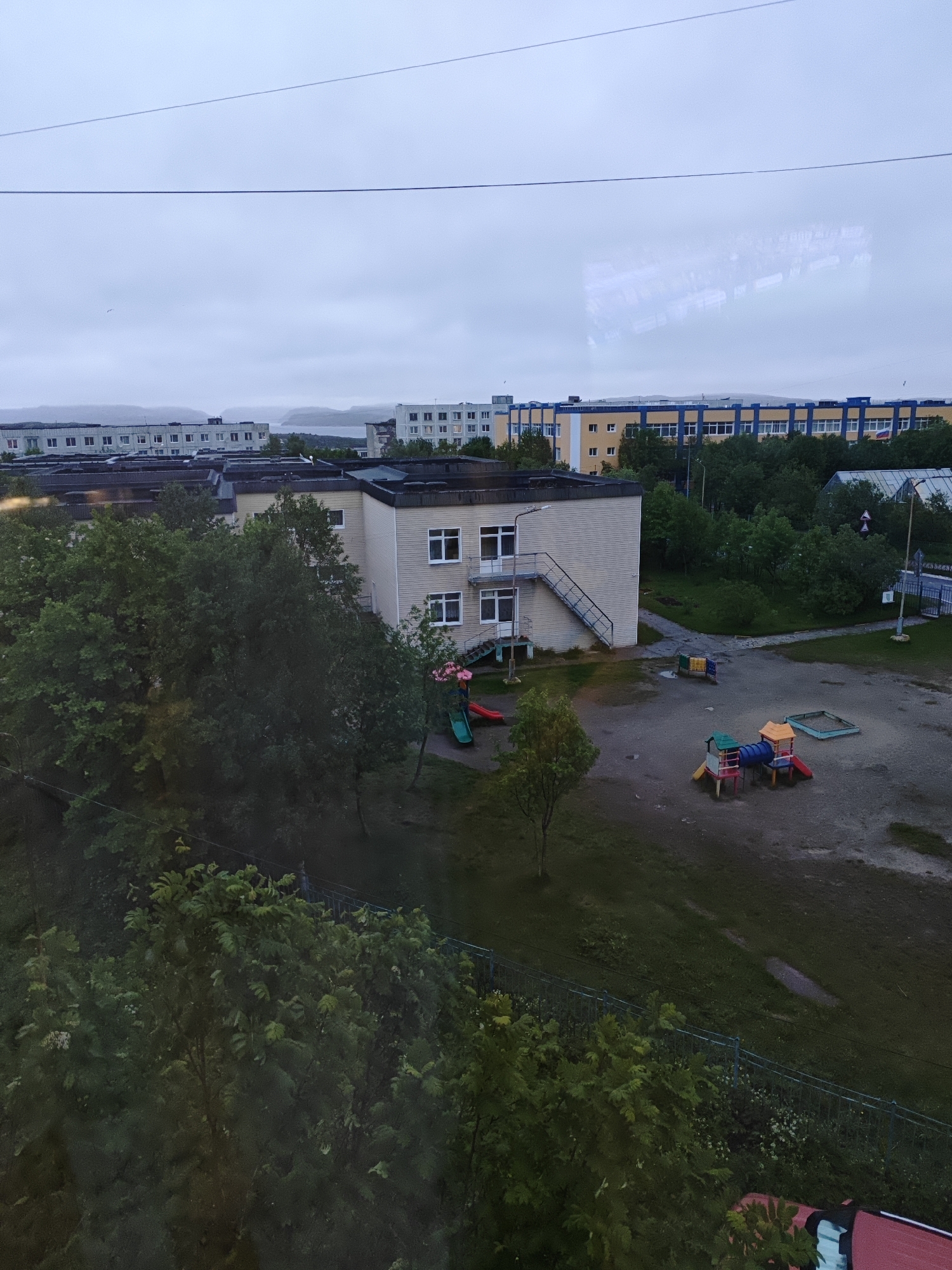 An outdoor, high-angle, full shot captures a grey, overcast day in a residential area. In the foreground, a lush green tree is partially visible. Beyond it, a light-colored, low-rise building with white-framed windows and an external staircase stands beside a playground featuring a blue slide, a tunnel with red and yellow sections, and a sandbox. Further back, more trees and a darker-colored building with a light-colored facade and white-framed windows are visible. In the distance, several larger, multi-story buildings with uniform windows dominate the landscape, with a body of water and hills in the far background. The sky is a uniform grey, with a thin electrical wire stretched across the top of the frame.