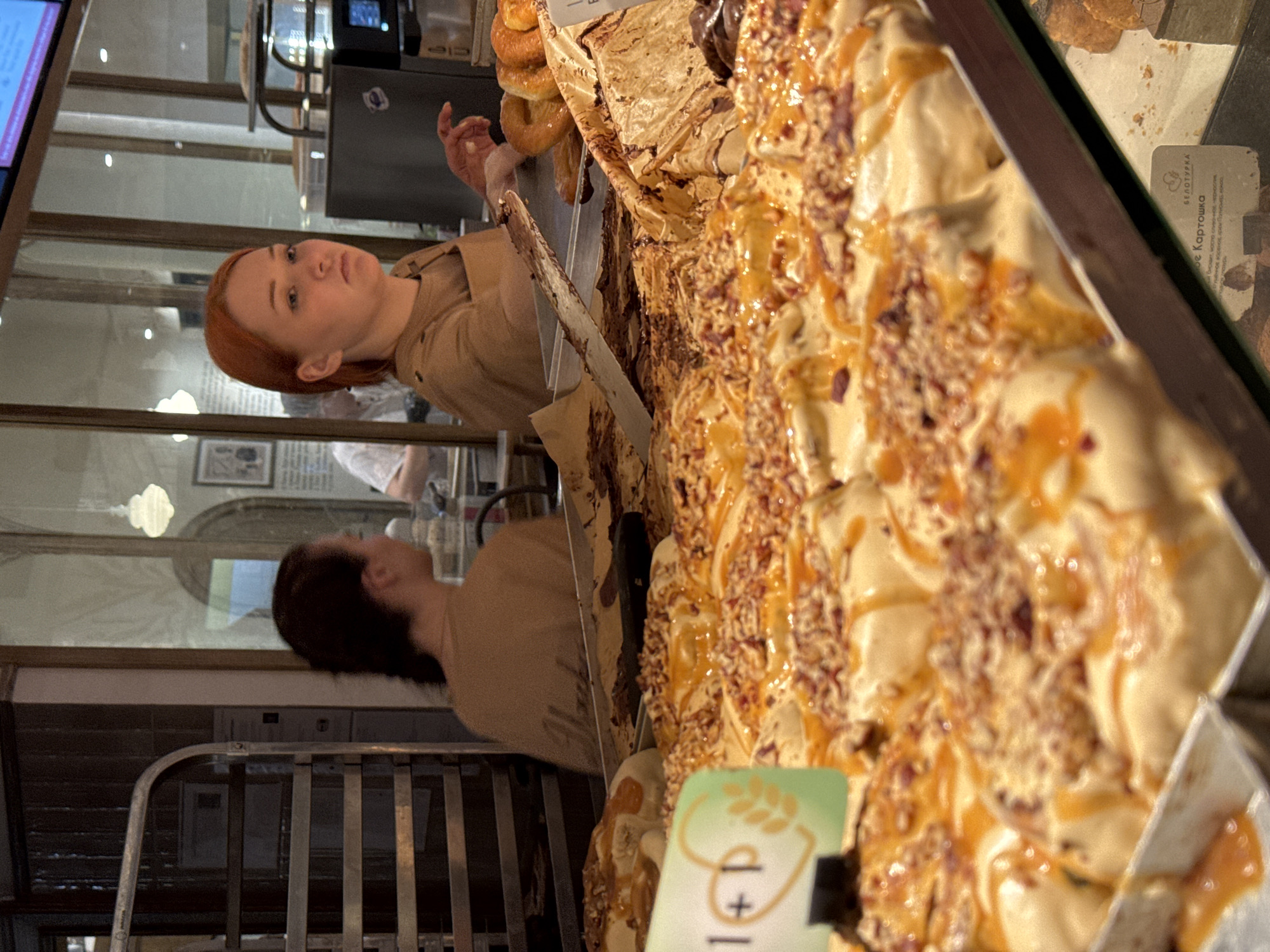 Here is a forensic description of the image:
**Scene:** An indoor scene possibly inside a bakery or food establishment. The focus is on a display case filled with pastries covered with a sweet caramel topping. There's a partially visible price tag with the number "1" on it.
**People:** There are two women reflected in the glass display case, with ginger hair and wearing light-brown clothing. One is closer to the pastries and possibly interacting with them.
**Objects:** A metal serving utensil is seen sticking out from between pastries. Various baked goods and pastries are behind the tray of caramel covered desert bars.
**Location:** The text on a label reads "Картошка", which translates to "potato". "Белотурка" is on the same label. The location is in Izhevsk, Russia.