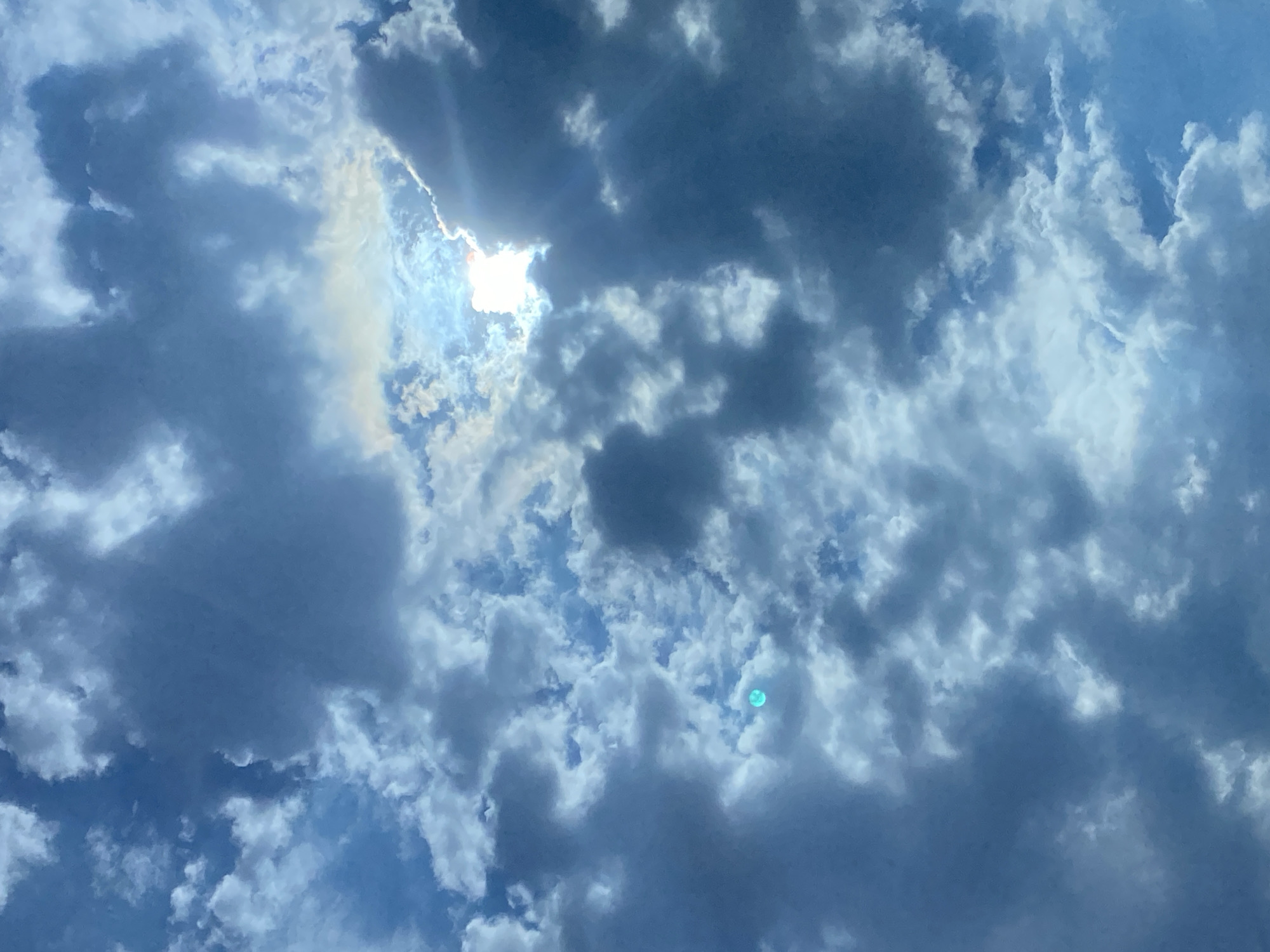 The image is a low-angle shot looking up at a cloudy sky. The sun is partially obscured by dark gray clouds, with rays of light breaking through. There are also patches of white, fluffy clouds scattered throughout the sky. In the center of the image, there is a bright, turquoise-colored circular anomaly, possibly a lens flare or reflection. The overall color palette is dominated by shades of blue and gray. The scene is entirely natural, with no man-made structures or elements visible. The location is described as Unknown, Unknown.