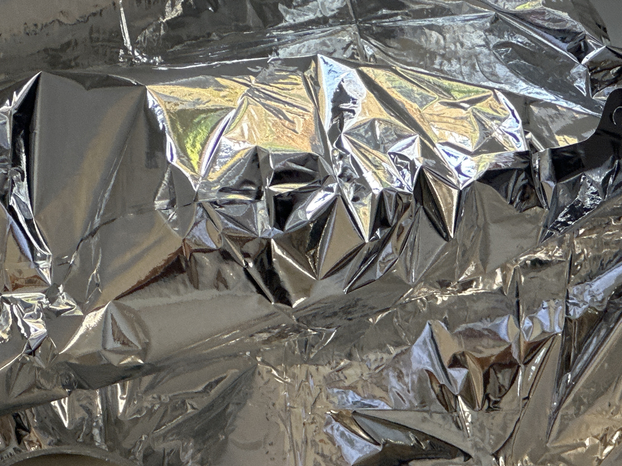The image displays a full-frame, close-up view of a heavily crumpled, reflective metallic silver material, consistent with a Mylar sheet or aluminum foil. The surface is characterized by numerous sharp creases, folds, and wrinkles, creating a complex, angular topography across the entire frame. Areas of intense specular reflection are interspersed with darker, less illuminated zones, indicating varied light absorption and reflection due to the material's irregular shape. Subtle color shifts, including yellowish, bluish, and greenish hues, are faintly visible within the reflections on various planes, suggesting ambient light sources or reflected objects of those colors. No individuals, discernible objects beyond the metallic material, or specific actions are present. The scene is static and abstract. The location is Wealden, United Kingdom.