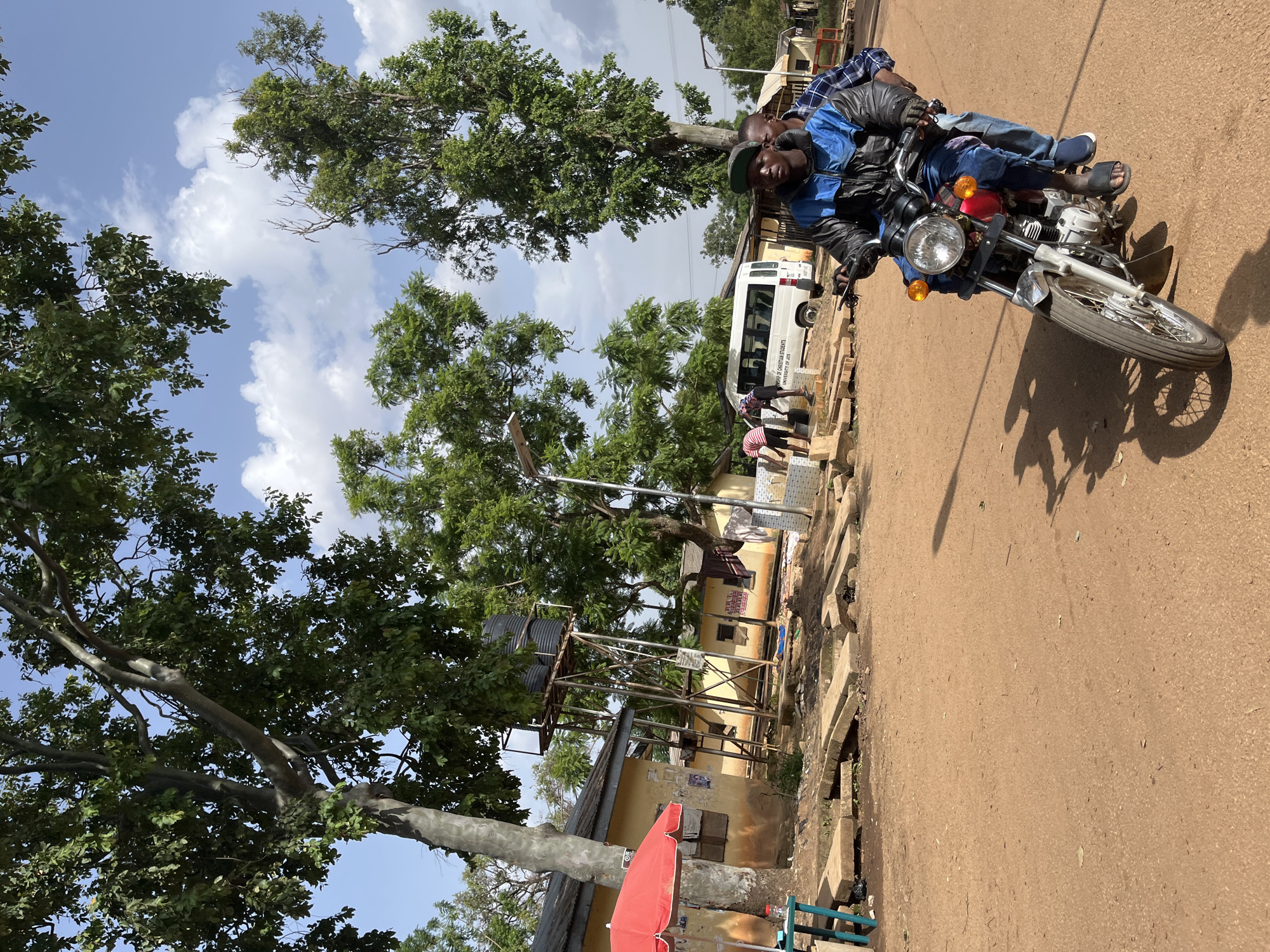In an outdoor scene in Jos, Nigeria, two individuals are riding a motorcycle on a dirt road. The driver is wearing a blue and black jacket and jeans, while the passenger behind him is wearing a plaid shirt. The motorcycle is silver with a red taillight. In the background, several yellow buildings are visible, some with corrugated metal roofs. A water tower with black tanks stands near one of the buildings. Trees with green foliage are scattered throughout the scene, and the sky is partly cloudy. A white van with text on its side is parked near the buildings. A red umbrella is open next to a turquoise table. The ground is covered in reddish-brown dirt, with scattered concrete slabs.