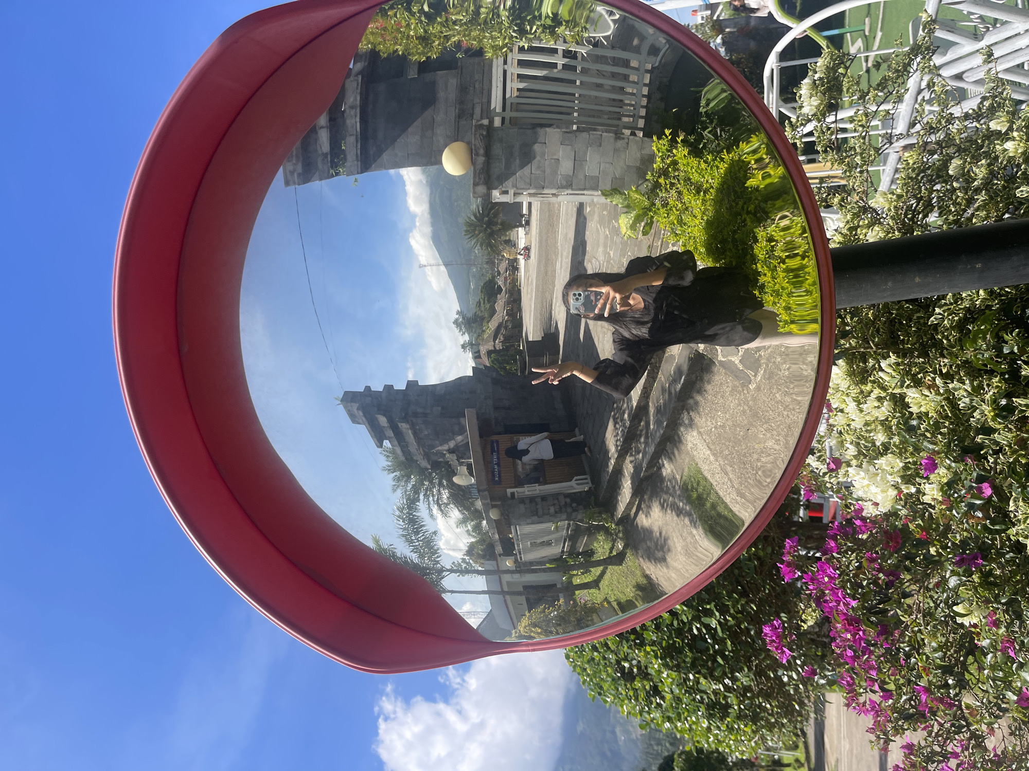 A person is reflected in a convex traffic mirror along a roadside in Kledung, Indonesia. The mirror's reflection shows a paved area, a building with stone walls and a gate, a distant mountain, and a cloudy sky. The person is holding a phone, seemingly taking a picture, and making a peace sign. The mirror itself has a red frame and is mounted on a black pole. Lush greenery, including flowering bushes with purple blooms, surrounds the pole.