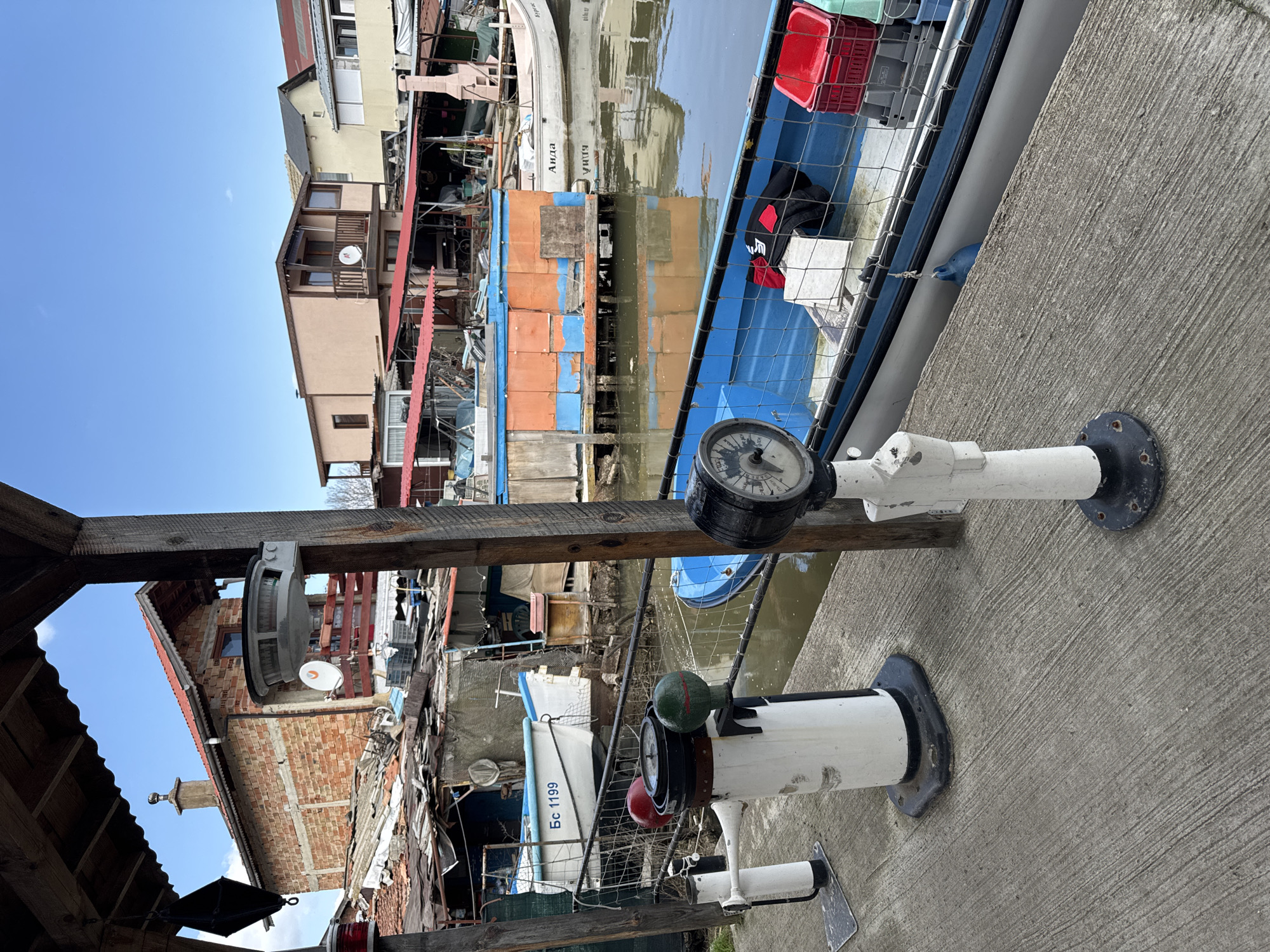 The image shows a waterfront scene in Burgas, Bulgaria. The camera is focused on a navigational instrument stand bolted to a concrete pier. The stand features a compass at the top, along with navigation lights (red and green) below. In the background, there's a waterway with several boats and boathouses. The structures appear somewhat ramshackle, constructed from various materials, including corrugated metal and wood. Buildings and houses are visible beyond the waterway, and the sky is a clear blue. The overall impression is one of a working waterfront area.