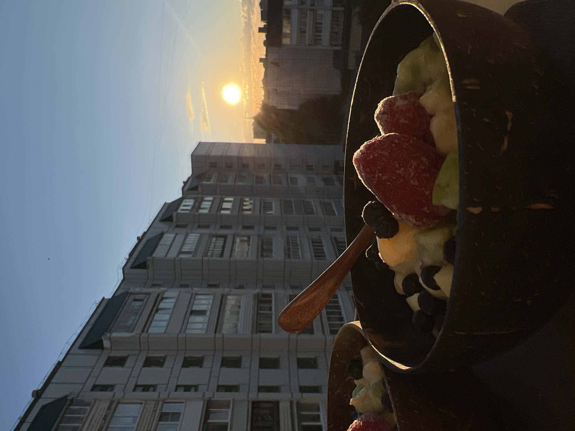 The image presents an outdoor scene at sunset, likely in Chaikovsky, Russia, featuring two bowls of fruit in the foreground against an urban backdrop. The photograph appears to be taken from an elevated position, such as a balcony or rooftop.
In the immediate foreground, two dark, rustic bowls, resembling polished coconut shells, are filled with a colorful array of fruits. The prominent bowl contains large, bright red strawberries, dark blueberries, and chunks of lighter-colored fruit, possibly banana or kiwi. The fruits have a slightly frosty appearance, suggesting they are chilled or frozen. A small, light brown wooden spoon is partially visible, resting in this bowl. A second, similar bowl is partially visible in the lower right corner, also containing fruit.
The background is dominated by multi-story residential buildings with numerous windows, characteristic of an urban or suburban environment. The sun, a brilliant golden disc, is low in the sky, illuminating the scene with a warm, soft glow, indicative of either sunrise or, more likely, sunset. The sky is clear and blue, with a few faint, wispy clouds near the horizon, tinged orange by the sun's light.
The overall impression is one of tranquility and enjoyment, capturing a moment of a refreshing meal or snack against a peaceful evening setting. No text is visible in the image.