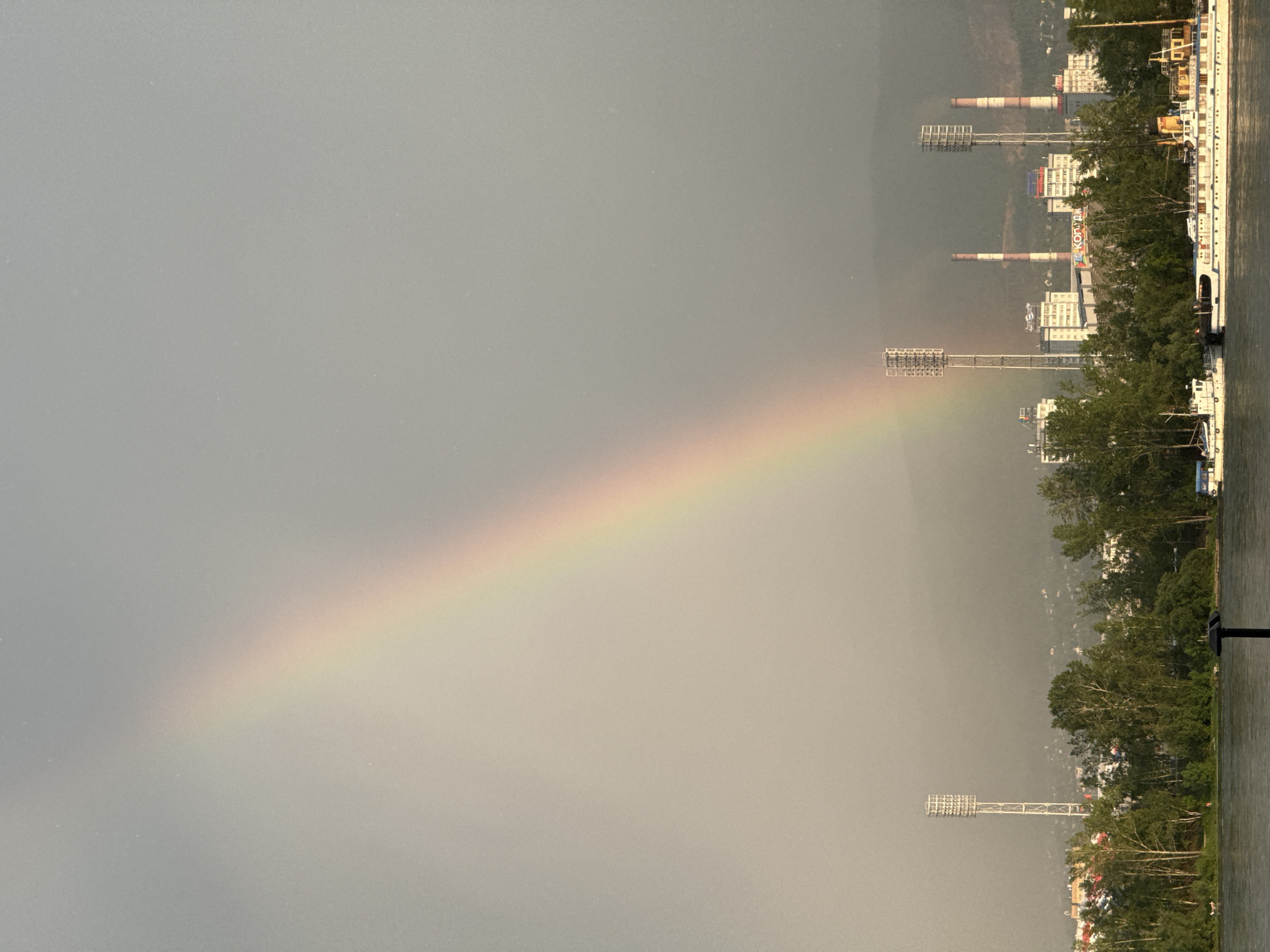 This is an outdoor, eye-level, medium shot photograph taken from the bank of a body of water, likely the Yenisei River, looking towards the cityscape of Krasnoyarsk, Russia. A prominent rainbow arcs across the hazy sky, its colors ranging from violet to red. In the mid-ground, several tall, white light towers stand at intervals, reminiscent of stadium lighting. Behind these towers, various buildings are visible, including industrial structures with tall smokestacks, some emitting faint smoke, and other multi-story buildings. Dense green trees line the waterfront in the foreground and middle ground, partially obscuring the buildings. Several boats are docked or moored on the water in the foreground, including a large, white passenger vessel. The sky is overcast, with the light suggesting either early morning or late afternoon. There are no visible people or animals in the frame, but the presence of boats and buildings indicates human activity and habitation. The overall atmosphere is serene, with the natural phenomenon of the rainbow contrasting with the industrial and urban elements of the city.