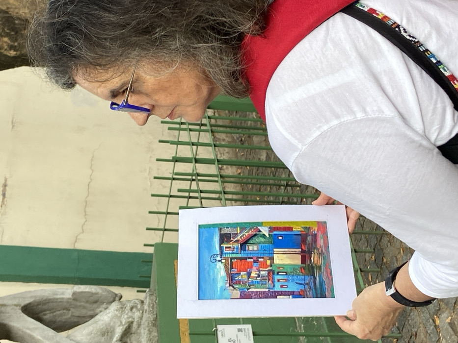 An older woman with gray hair and blue-framed glasses closely examines a framed painting. The painting depicts colorful buildings, predominantly blue and red, in an abstract style. The woman wears a white shirt with a red collar and a decorative strap. A green metal fence and a light-colored building are visible in the background. A stone structure is visible on the left.