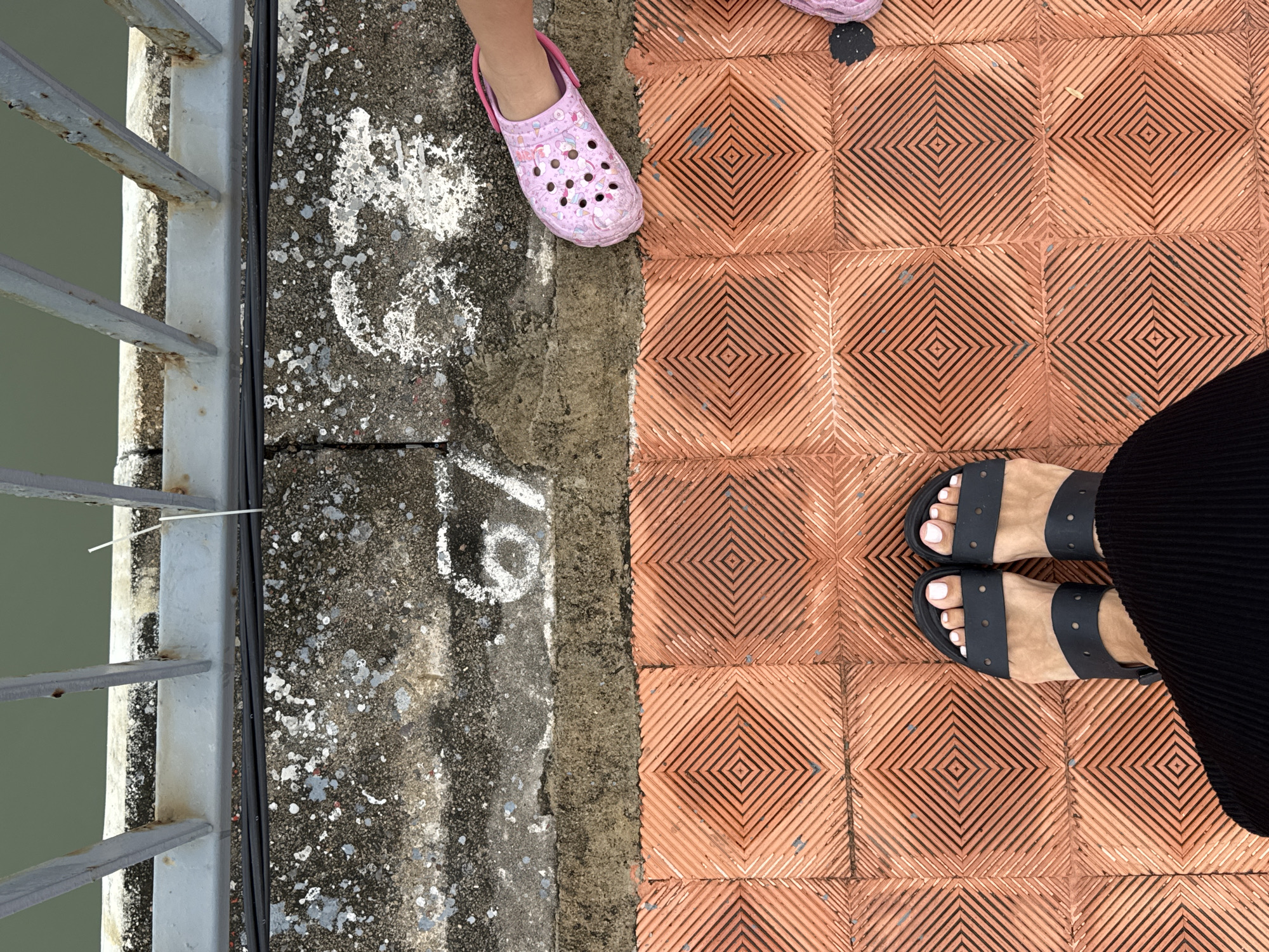 An eye-level overhead shot shows a concrete walkway with patterned reddish-brown tiles to the right and rough, mottled gray concrete to the left, alongside a weathered metal railing. Two pairs of feet are visible: a child's foot wearing a pink cartoon-printed Croc is positioned partially on the gray concrete and partially on the patterned tiles, and an adult's feet wearing black sandals stand on the patterned tiles. A section of a black pleated garment is also visible near the adult's feet. The location is Bac Nha Trang Ward, Vietnam. A white cable tie is visible on the metal railing, suggesting a makeshift repair or attachment.
