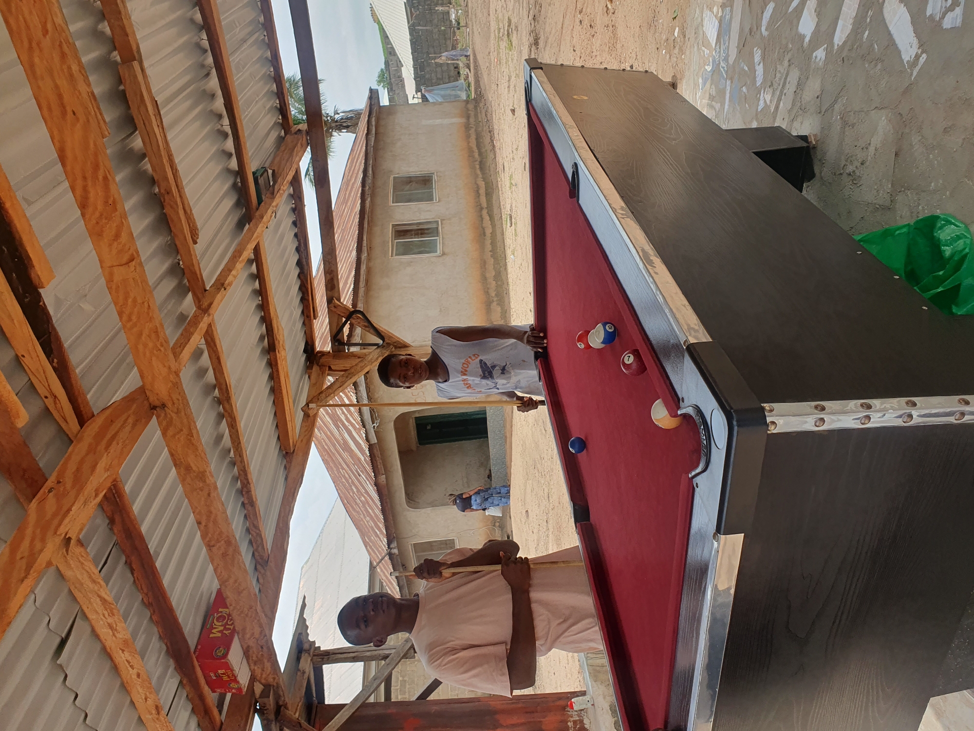 Two young boys are standing at either end of a pool table, holding cues. The pool table has a red felt surface and is situated outdoors under a wooden-framed roof with corrugated metal sheeting. In the background, there is a modest, light-colored building with windows and a tiled roof. The ground is a mix of dirt and concrete. A green plastic bag is visible near the leg of the pool table. The scene suggests a casual recreational setting in Lokoja, Nigeria.