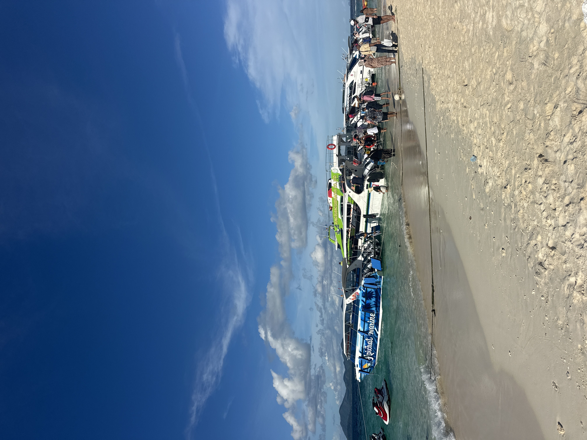 This is a horizontal, eye-level, medium shot of a sandy beach lined with several boats on a bright, sunny day. The sky is a vivid blue with scattered white clouds. The sand is light brown and appears dry and granular. Several people are visible, some standing on the beach and others on or near the boats. They are dressed in casual clothing, suggesting a tropical or vacation setting.
The boats are of various sizes and colors, predominantly white and green, with some appearing to be speedboats or tour boats. They are docked along the shoreline, and some have people boarding or disembarking. Two jet skis are visible in the shallow water near the shore. The water is clear and turquoise, with gentle waves lapping at the sand. In the background, the silhouette of a mountainous landmass can be seen against the sky.
Visible text on one of the boats reads "PICHAT MARINE". Another boat has text that appears to read "PHUNGUANG SADACHA CO., LTD.". The overall scene depicts a lively tourist area with recreational watercraft, likely a popular spot for beach activities and boat tours. The bright sunlight and clear sky indicate pleasant weather, and the presence of people suggests a daytime activity. The location is identified as Phru Nai, Thailand.