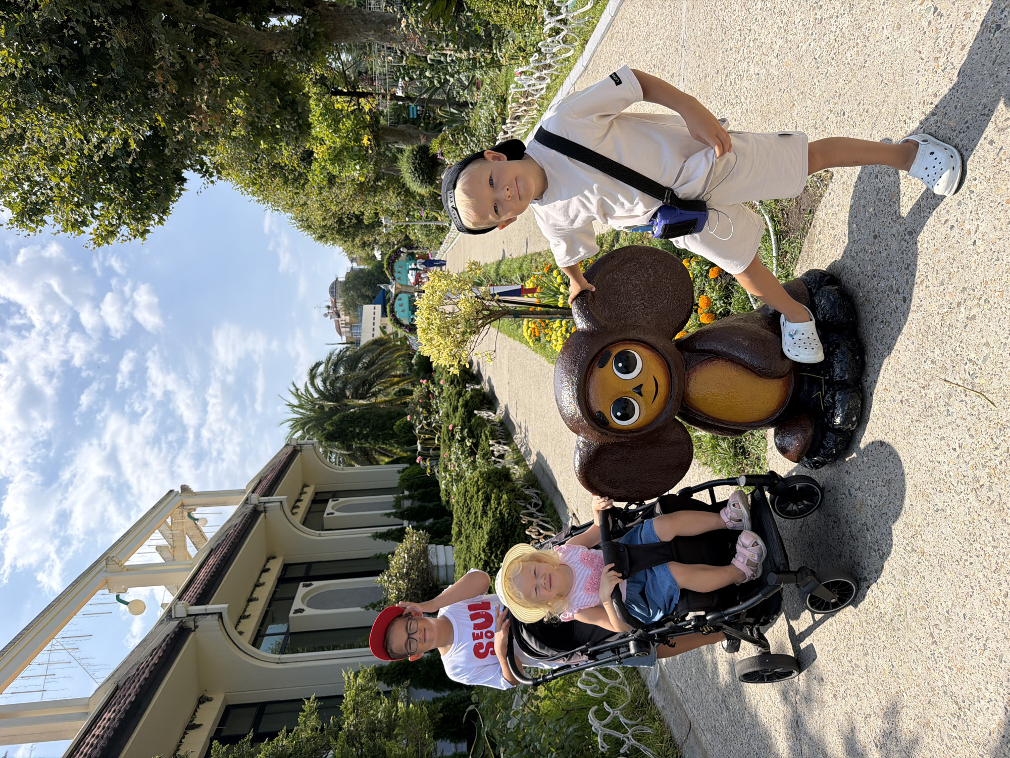 In Sochi, Russia, a boy wearing a black cap and a white t-shirt stands to the right of a statue of Cheburashka, a beloved cartoon character. He has a blue device with white earbuds hanging around his neck. A young girl in a straw hat and a pink-striped dress sits in a stroller to the left, and a boy wearing glasses and a red cap is behind the stroller, touching his hat. The children are positioned on a paved walkway lined with green bushes, trees, and white railings. The background features a white building with arched windows and a blue sky with white clouds.